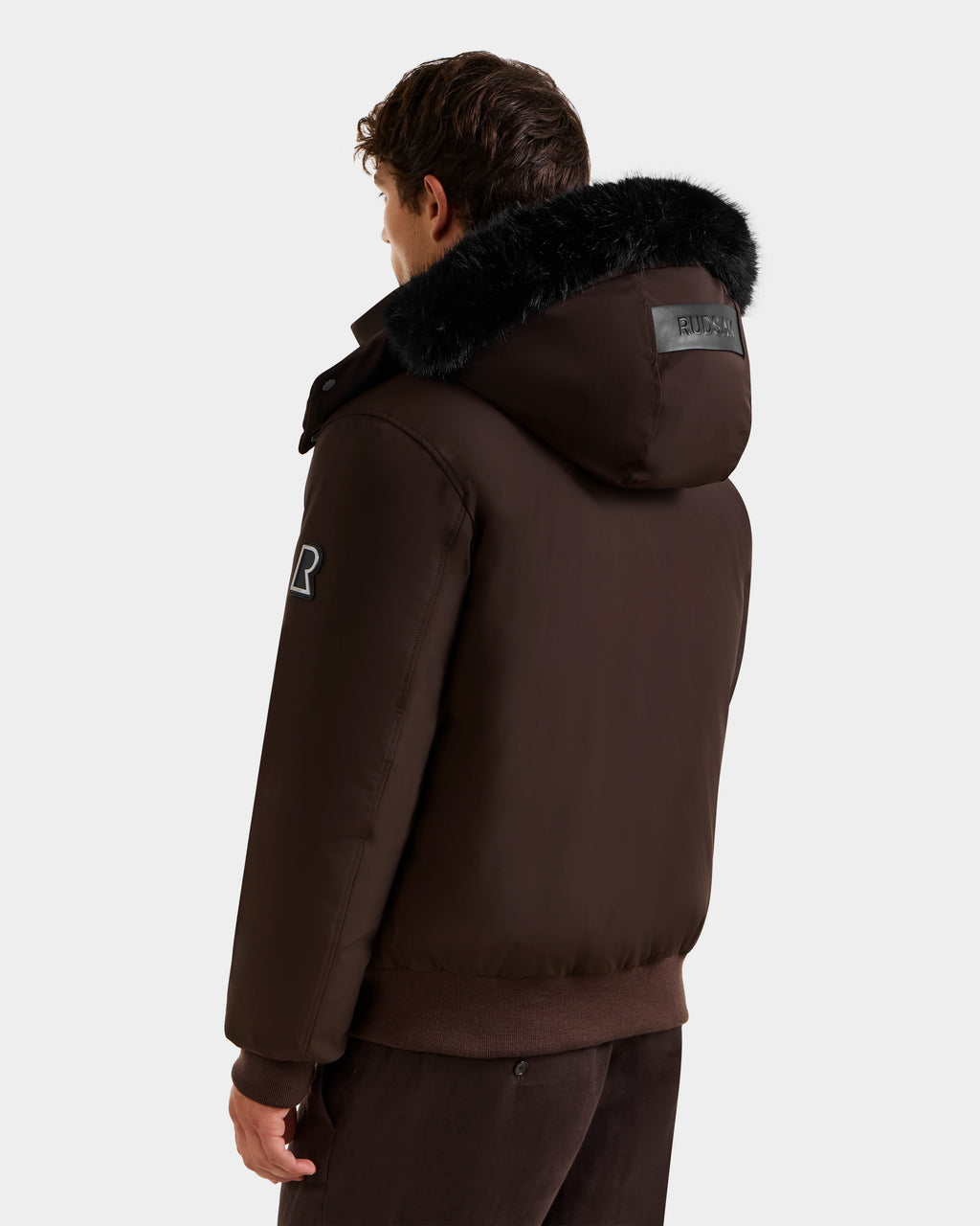 A person is shown from the back wearing the Rudsak JEROME - 6125514 Espresso men’s down bomber with black fur trim, a patch on the sleeve, and a rectangular black label on the hood, set against a plain light background.
