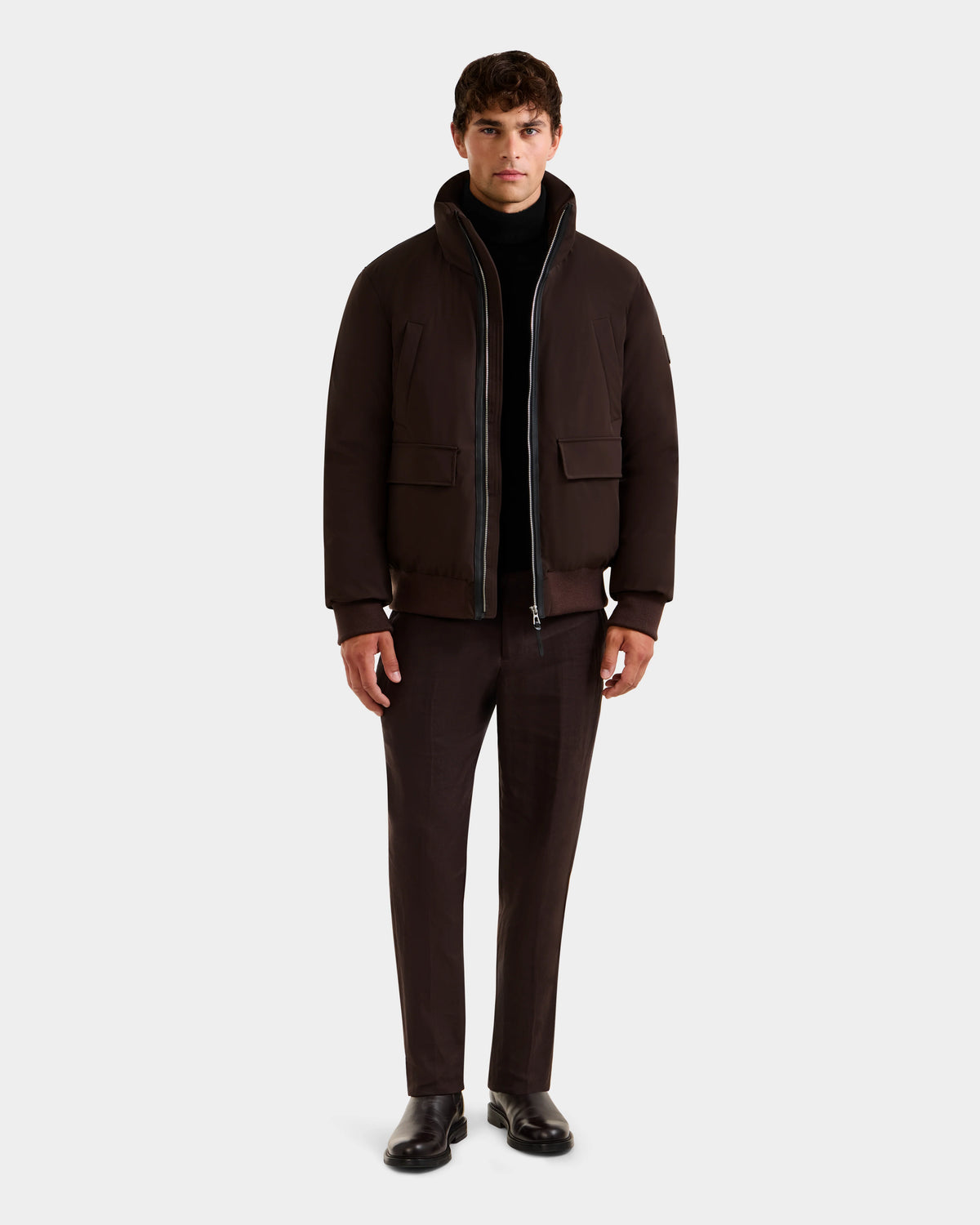 A man faces forward wearing the Rudsak JEROME - 6125514 Espresso men’s down bomber jacket, a black turtleneck, matching brown trousers, and black dress shoes against a plain light background.