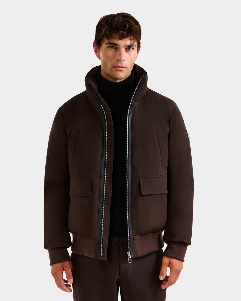 A man wears the JEROME - 6125514 Espresso down bomber jacket by Rudsak, featuring a high collar and front zipper, layered over a black turtleneck. He stands facing forward against a plain, light background.