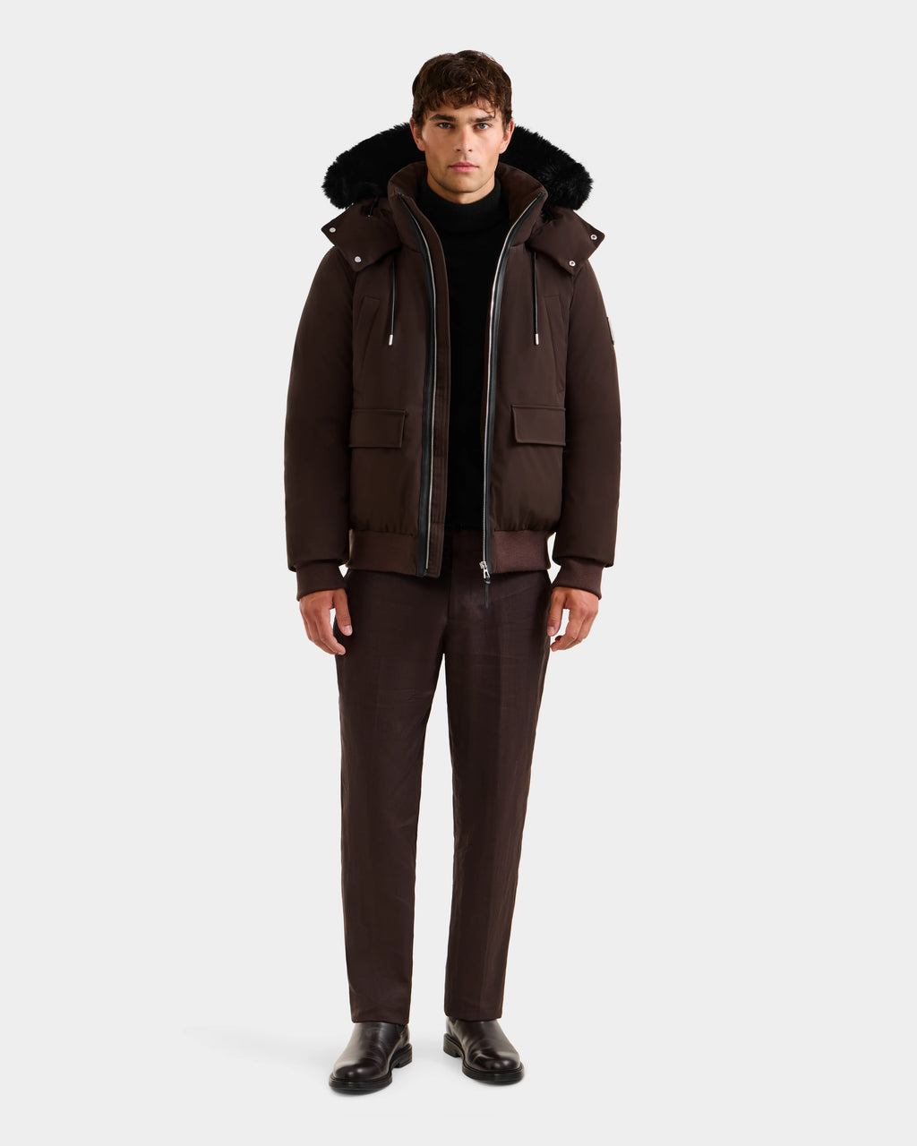 A man faces forward against a white background, wearing the Rudsak JEROME - 6125514 Espresso men’s down bomber jacket with large front pockets, dark trousers, a black turtleneck, and black shoes.