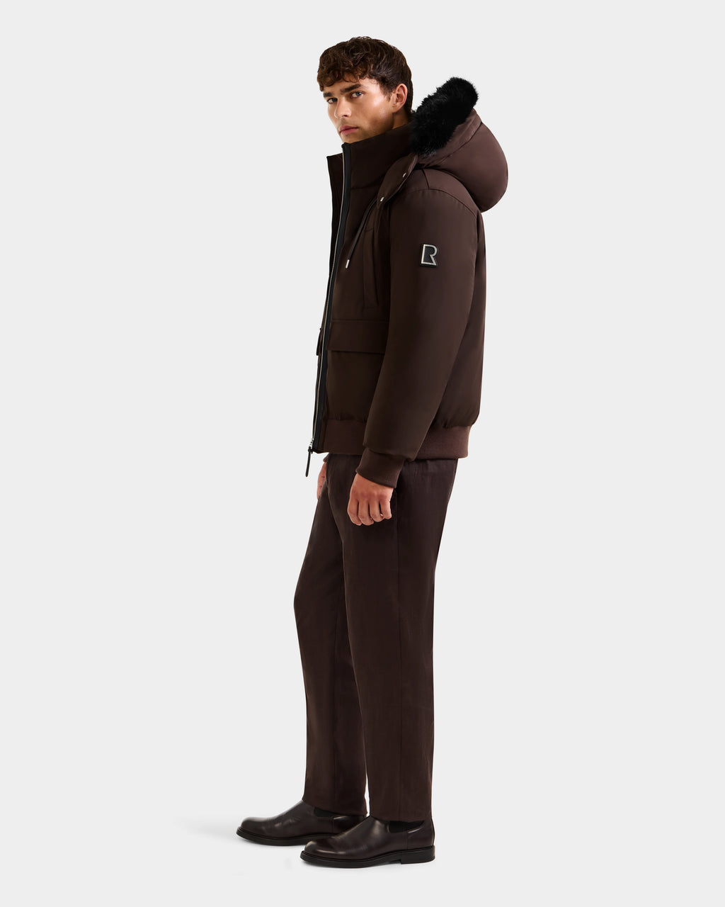A man stands sideways wearing the Rudsak JEROME - 6125514 Espresso men’s down bomber jacket with black fur trim, brown pants, and black shoes against a plain white background.