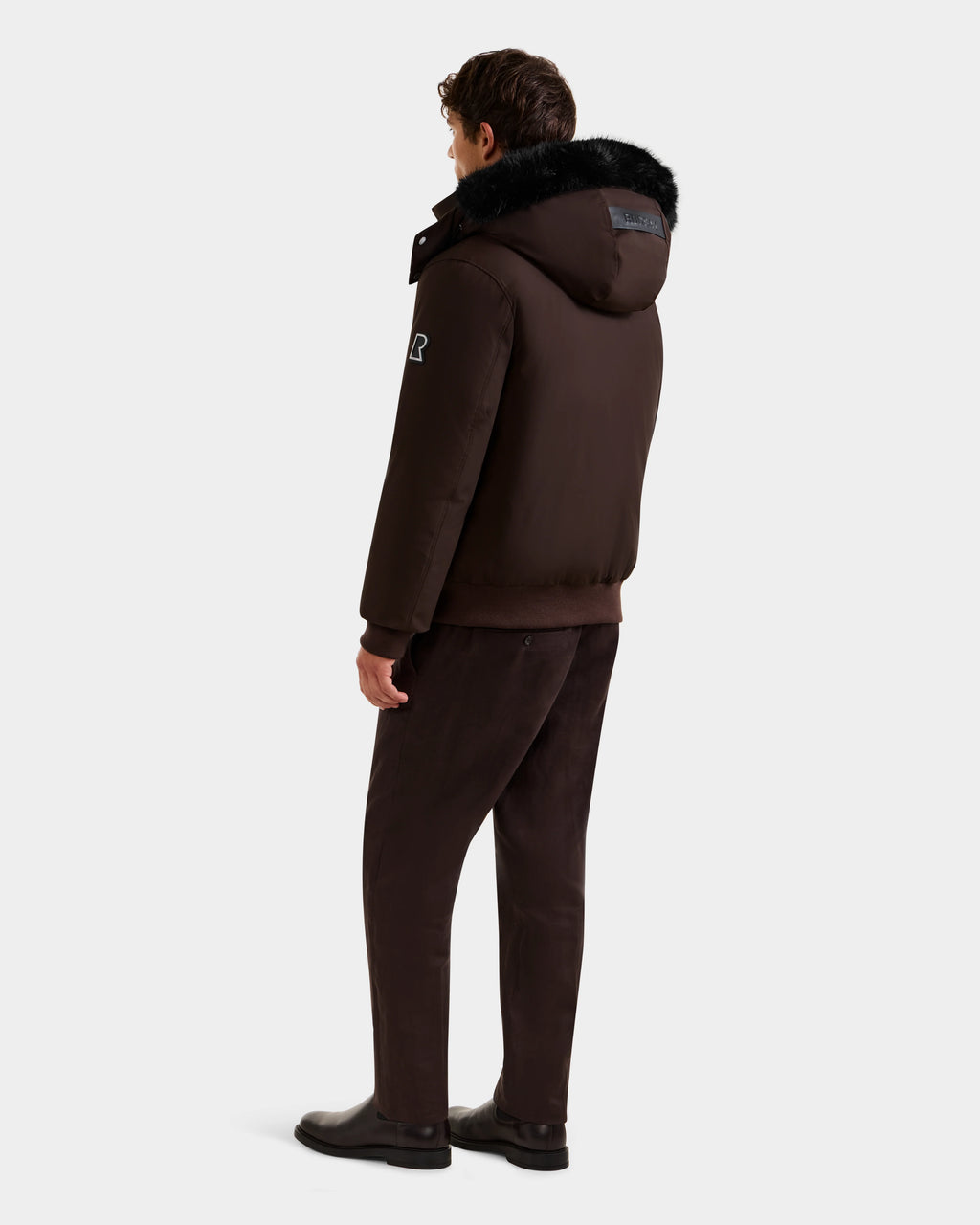 A man faces away wearing the Rudsak JEROME - 6125514 Espresso mens down bomber jacket with black fur trim, paired with matching dark brown pants and black shoes against a plain light background.
