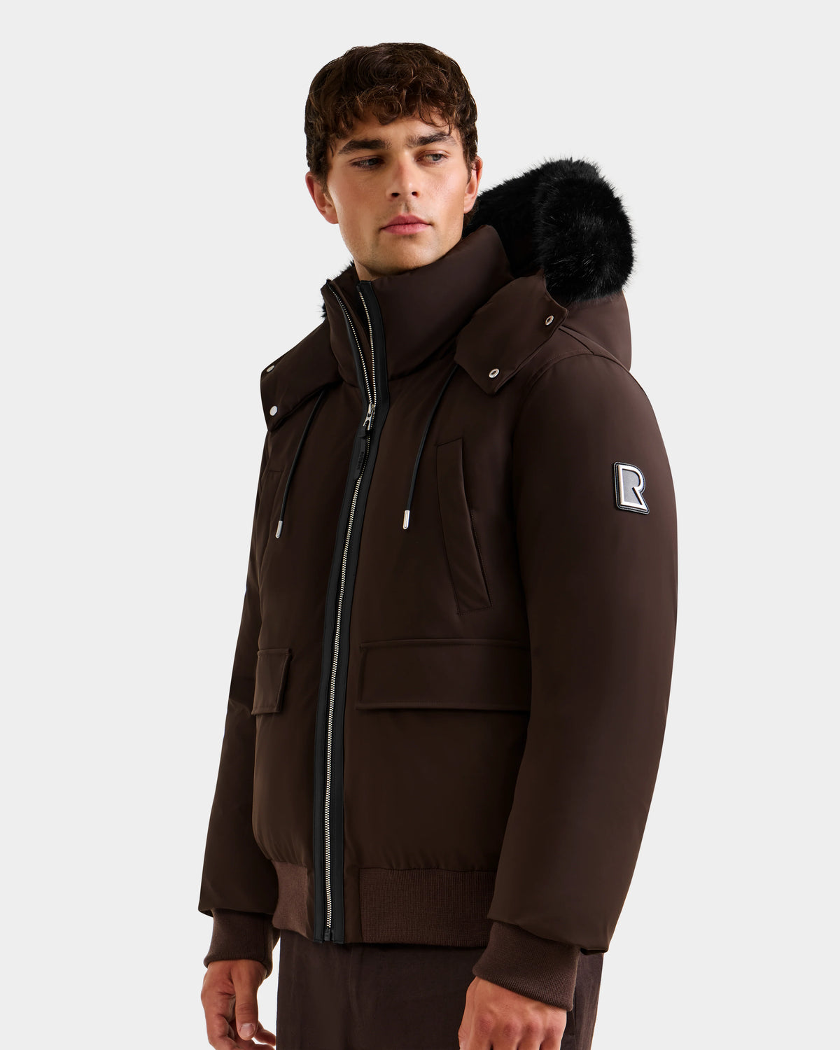 A young man stands against a plain background wearing the Rudsak JEROME - 6125514 Espresso, a men’s down bomber jacket with a black fur-trimmed hood, zippered pockets, RDS-certified down, and an R patch on the upper sleeve.