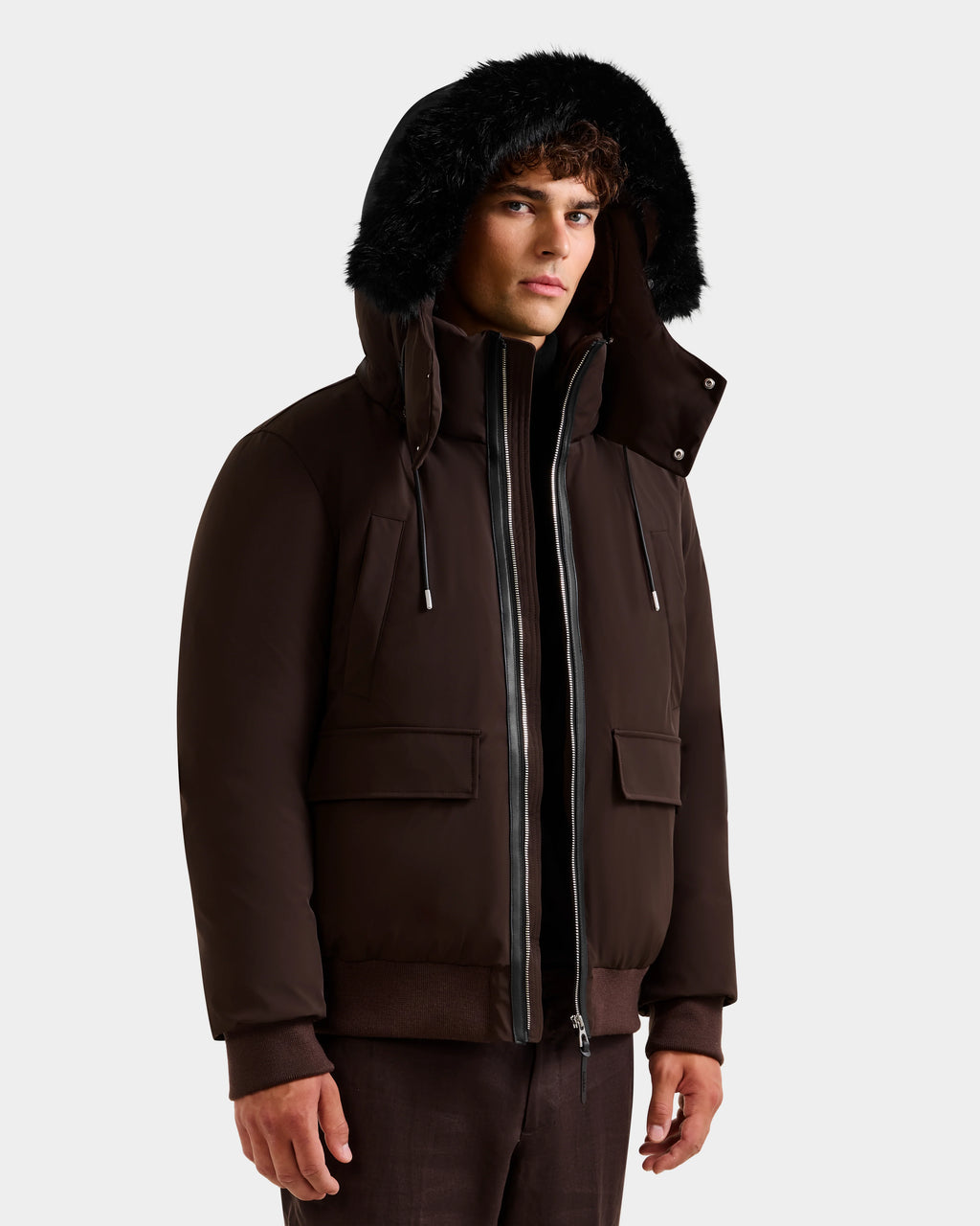 A man wears the Rudsak JEROME - 6125514 Espresso down bomber jacket with black fur trim, front zipper, and flap pockets over brown pants—an ideal winter choice for warmth and style against a light background.