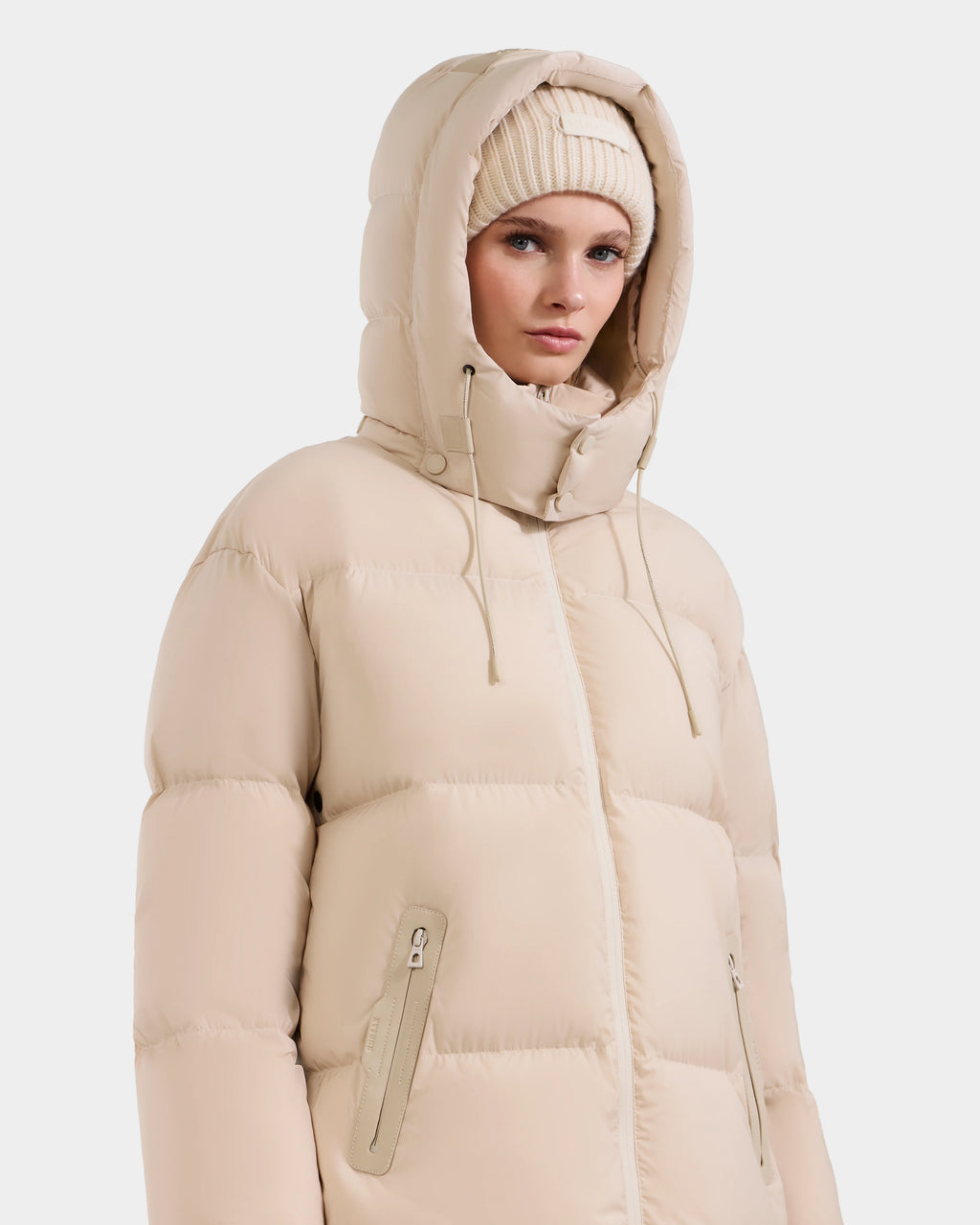 A woman wears the Rudsak KARLEE L - 8125682 Vanilla jacket, a beige puffer with vegan leather trims, drawstring hood, and zippered pockets, paired with a beige knit beanie, standing against a plain white background.