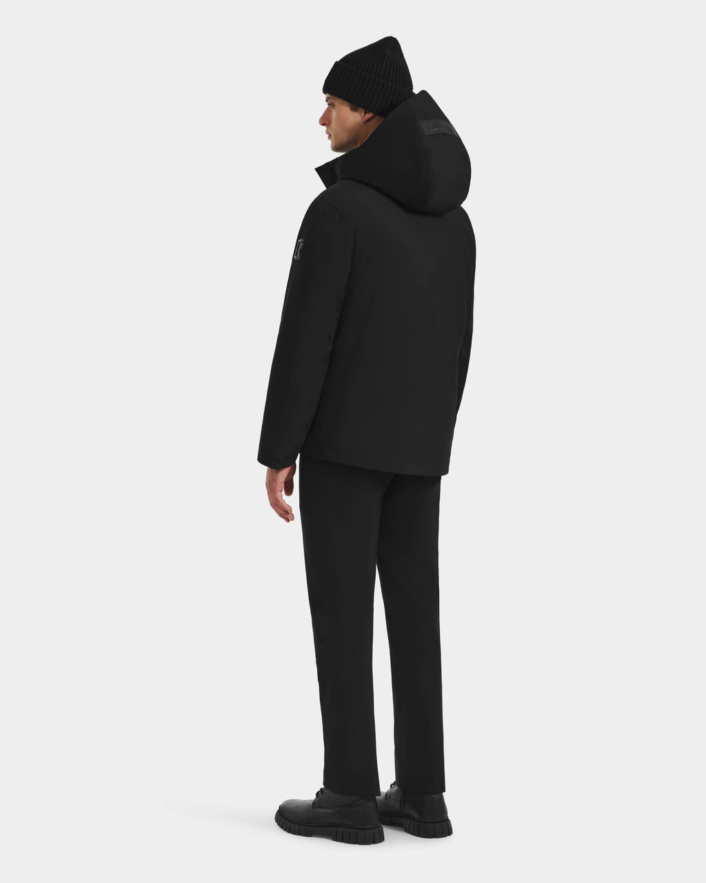 A man faces away, dressed in the Rudsak LAZIO - 6125527 Black down and feather fill men’s parka, with black beanie, pants, and shoes, set against a plain white background.