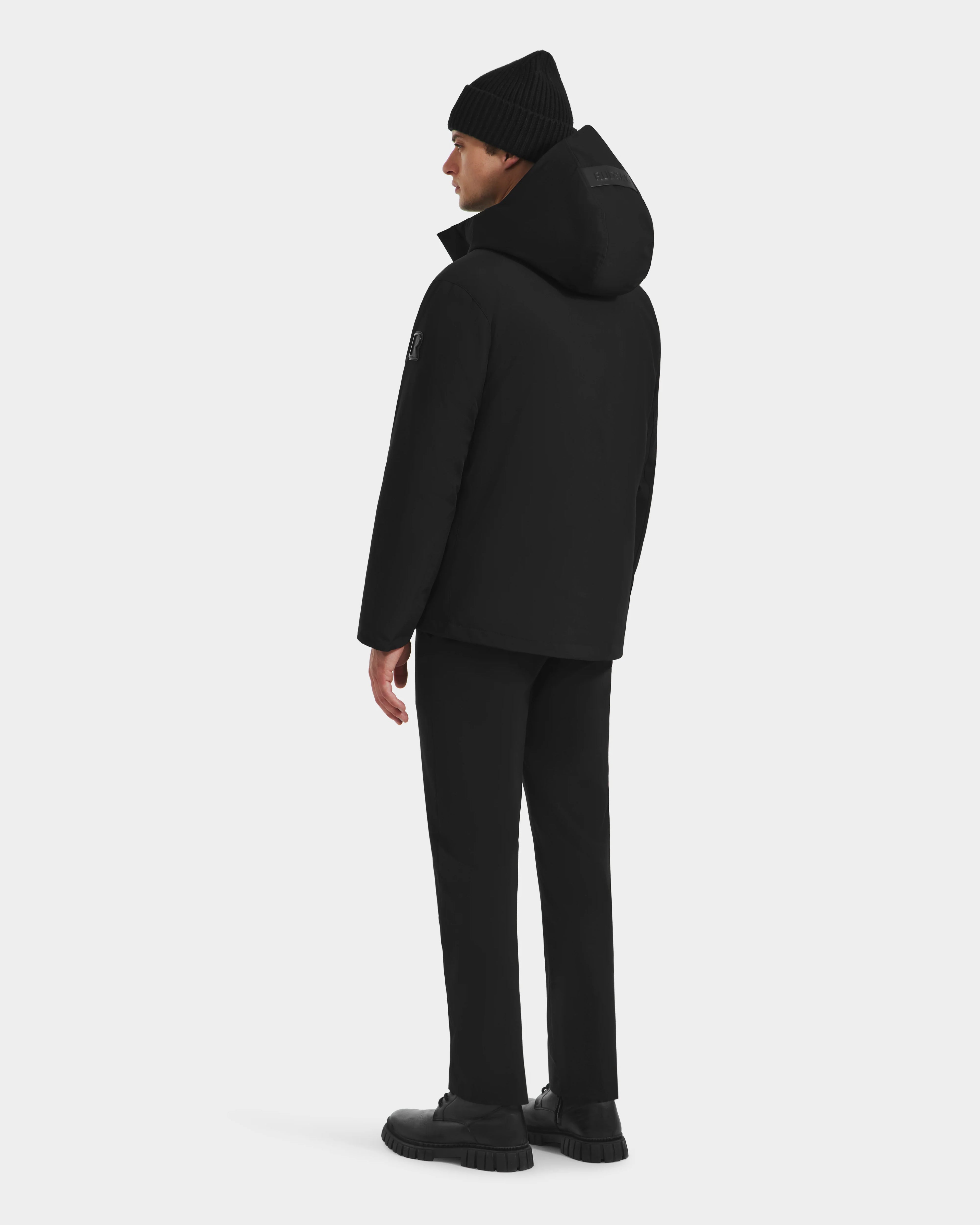 A man faces away, dressed in the Rudsak LAZIO - 6125527 Black down and feather fill men’s parka, with black beanie, pants, and shoes, set against a plain white background.