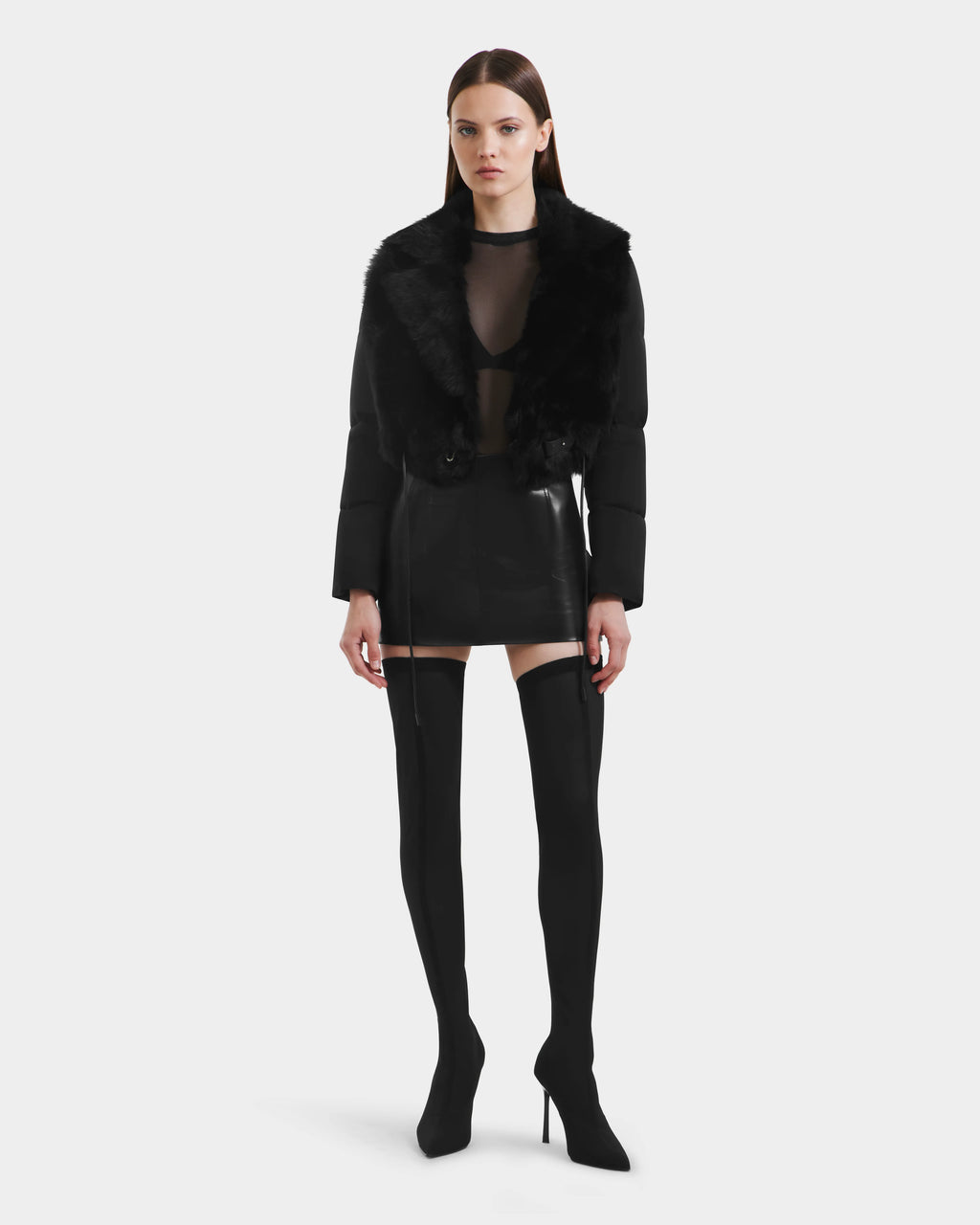 A woman faces forward against a white background, wearing the Rudsak LILAH - 8125509 Black shearling lamb jacket with quilted puffer sleeves, a sheer black top, black mini skirt, and over-the-knee heeled boots.