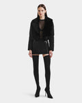 A woman faces forward against a white background, wearing the Rudsak LILAH - 8125509 Black shearling lamb jacket with quilted puffer sleeves, a sheer black top, black mini skirt, and over-the-knee heeled boots.