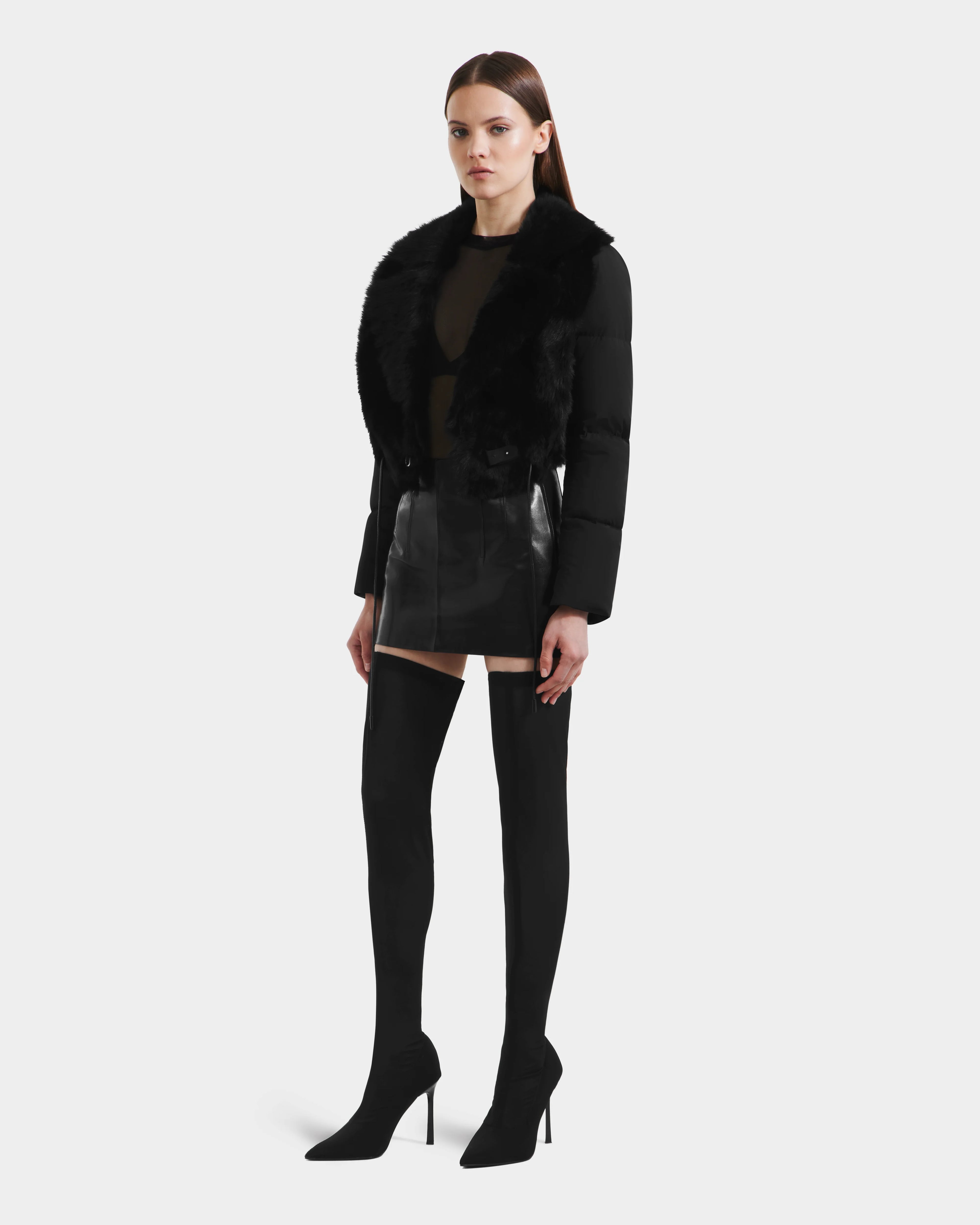 A woman poses against a plain background wearing the Rudsak LILAH - 8125509 Black women’s cropped jacket with faux fur collar, paired with a sheer black top, shiny black mini skirt, and thigh-high heeled boots.