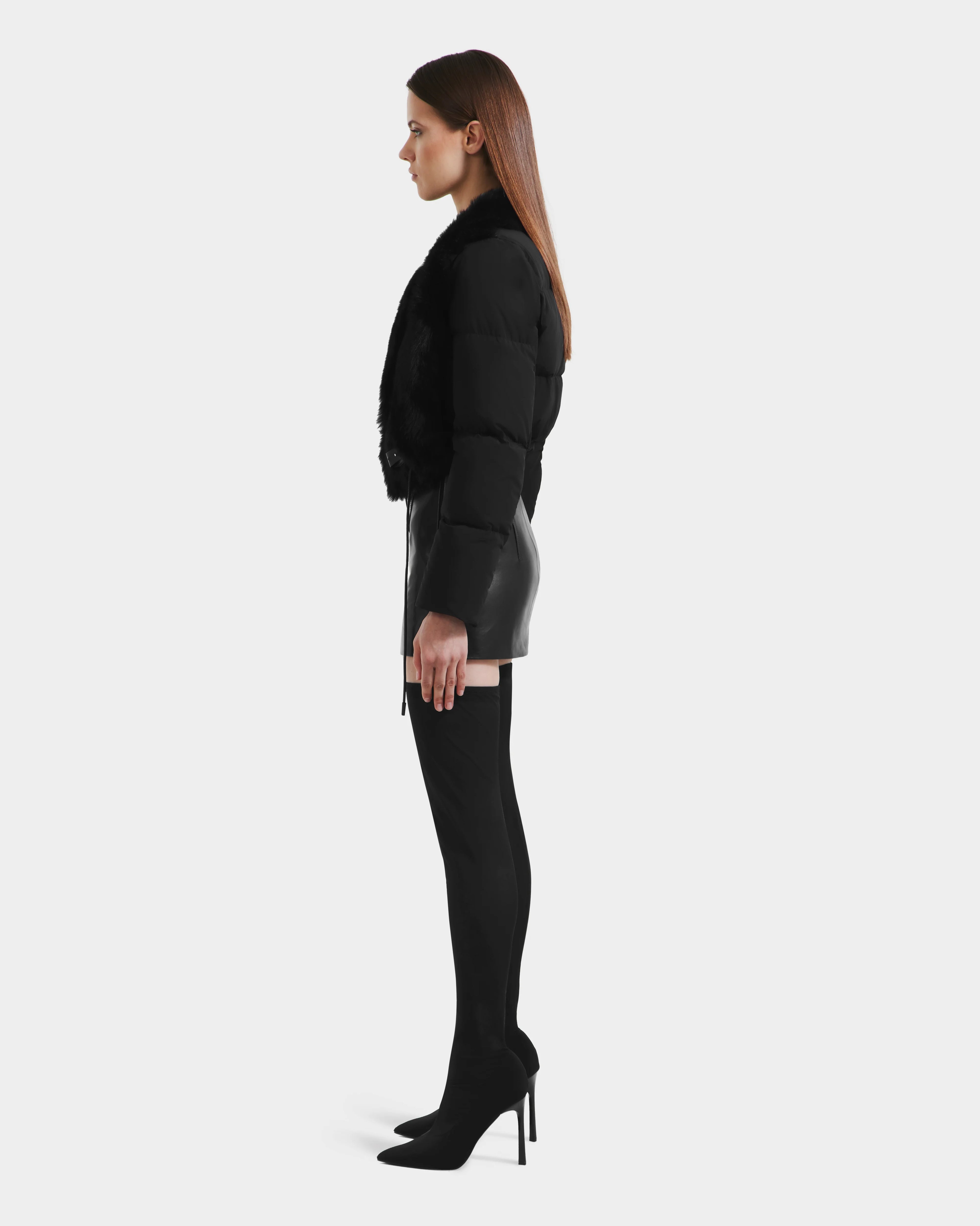 A woman stands in profile against a plain background, wearing the Rudsak LILAH - 8125509 Black women’s cropped jacket with quilted puffer sleeves and fur trim, styled with a black leather mini skirt and thigh-high heeled boots.