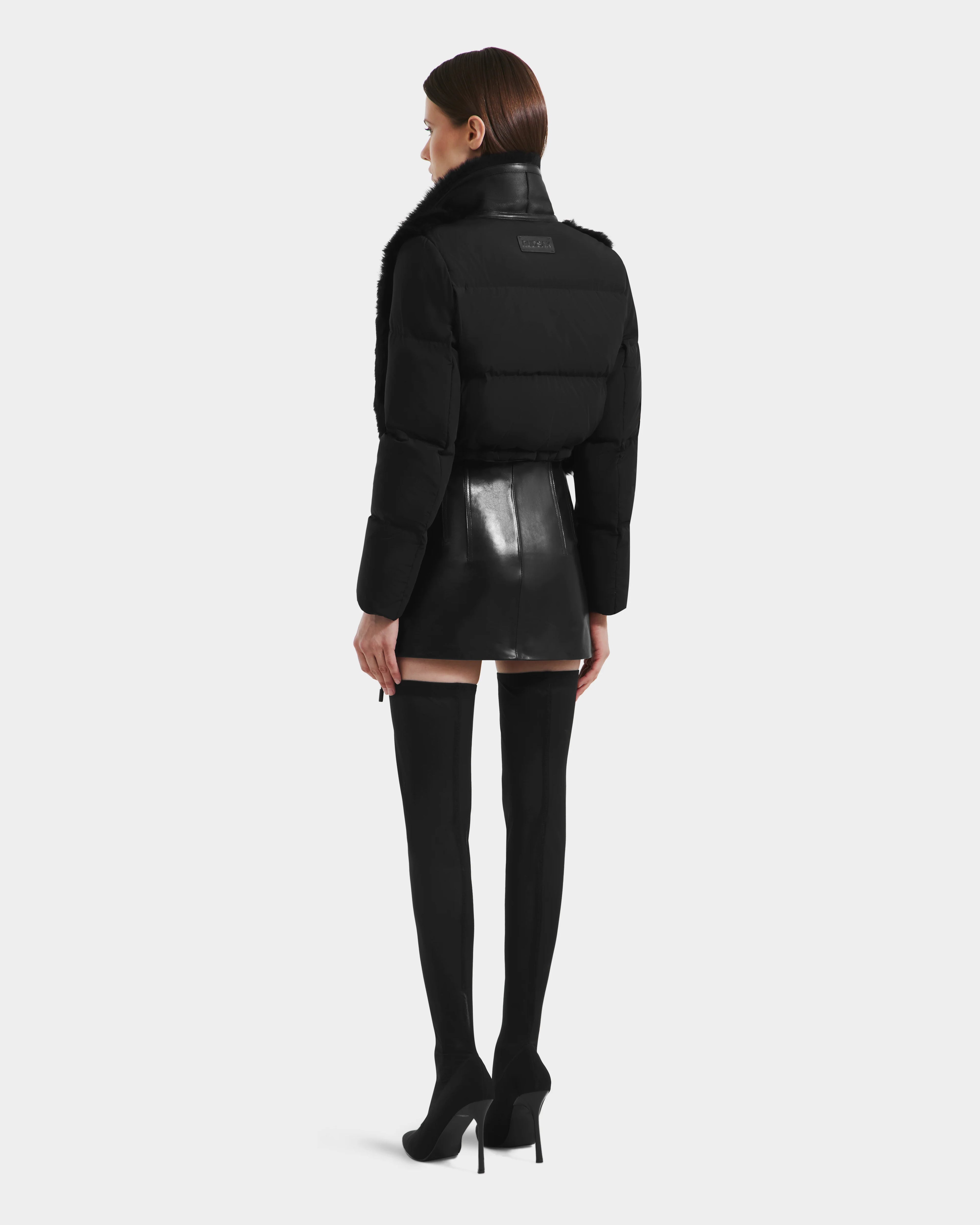 A woman stands facing away, wearing the Rudsak LILAH - 8125509 Black cropped jacket with quilted puffer sleeves, paired with a black mini skirt and thigh-high heeled boots. Her straight brown hair contrasts with the plain white background.