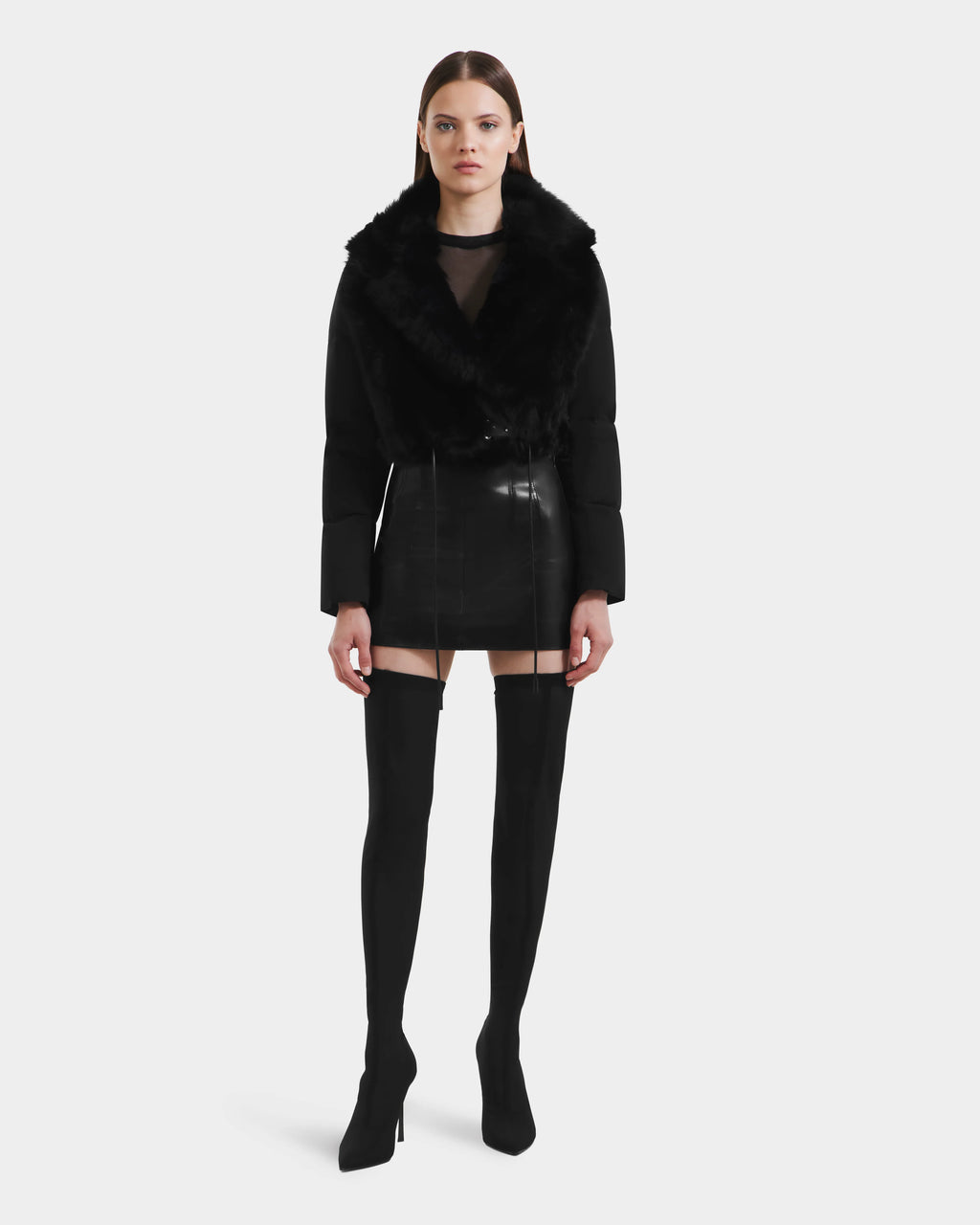 A woman faces forward in a Rudsak LILAH - 8125509 Black shearling lamb jacket with quilted puffer sleeves, worn over a sheer top, paired with a black leather mini skirt and black thigh-high heeled boots against a plain white background.