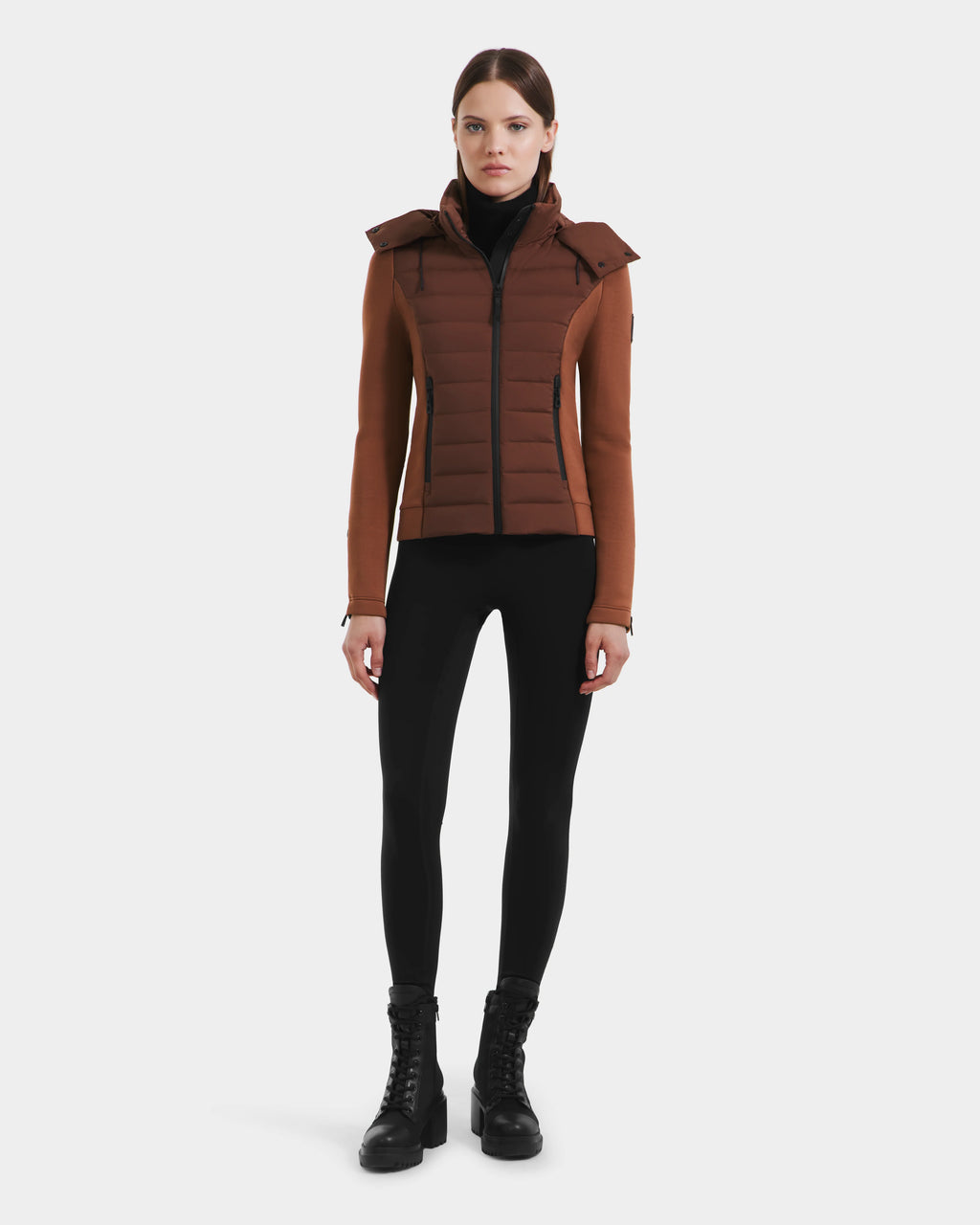 A woman faces forward against a white background, wearing the Rudsak MABS - 8125055 Rust puffer jacket with a hood, paired with a black turtleneck, black leggings, and black lace-up boots.