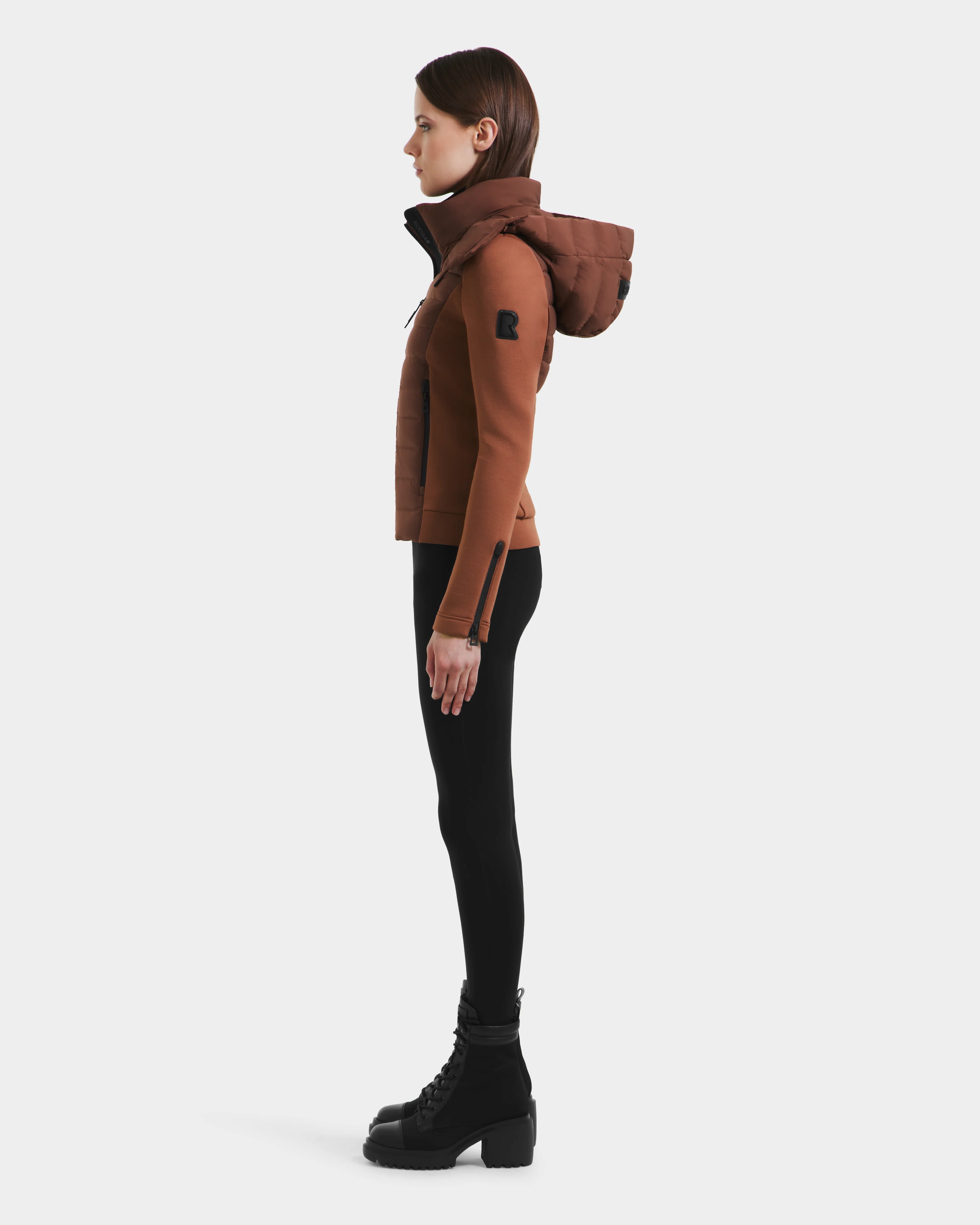 A woman stands in profile wearing the Rudsak MABS - 8125055 Rust lightweight activewear jacket with black leggings and lace-up boots against a plain white background.