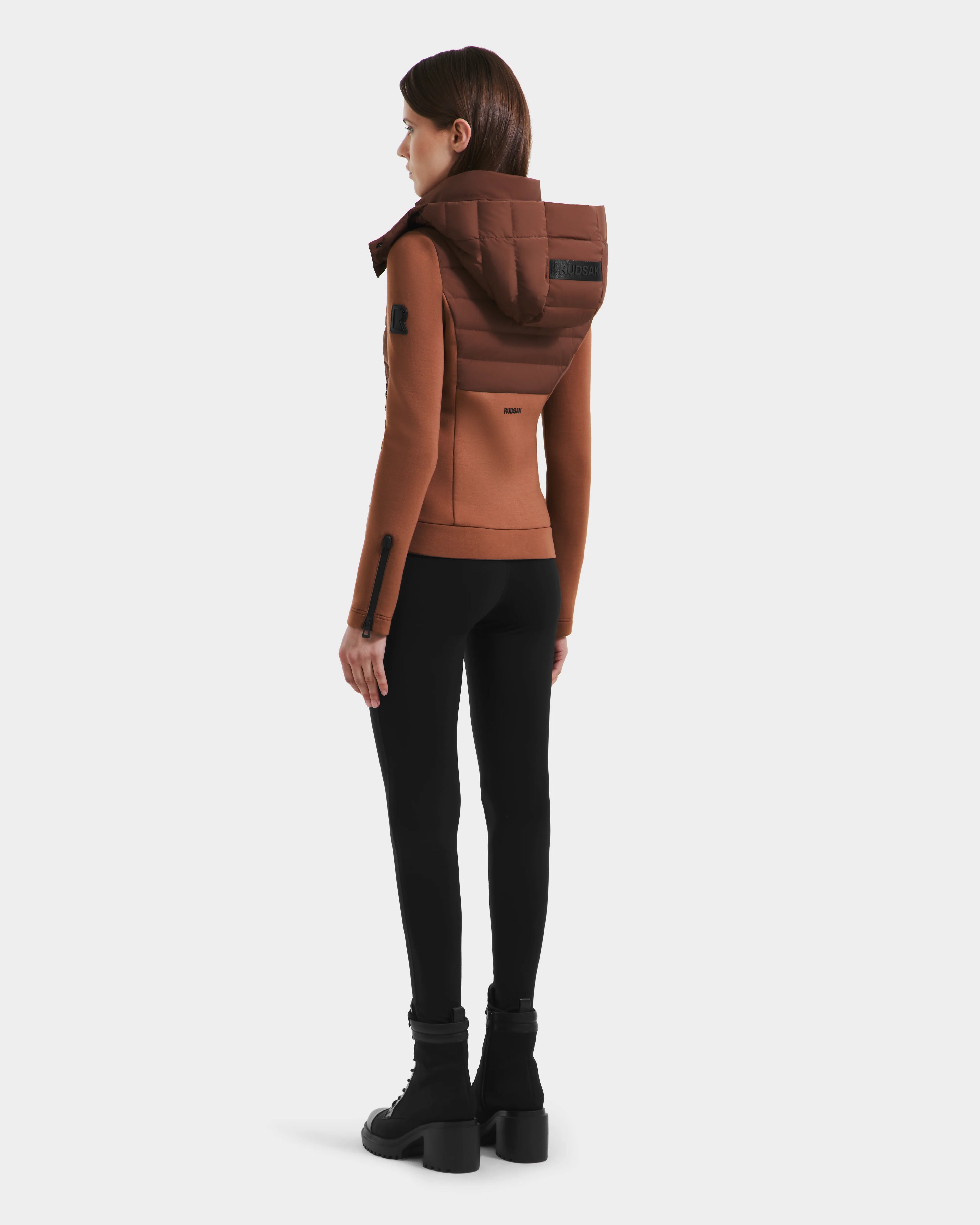 A person faces away, wearing the Rudsak MABS - 8125055 Rust lightweight activewear jacket with a large hood, paired with black leggings and boots, set against a plain light background.