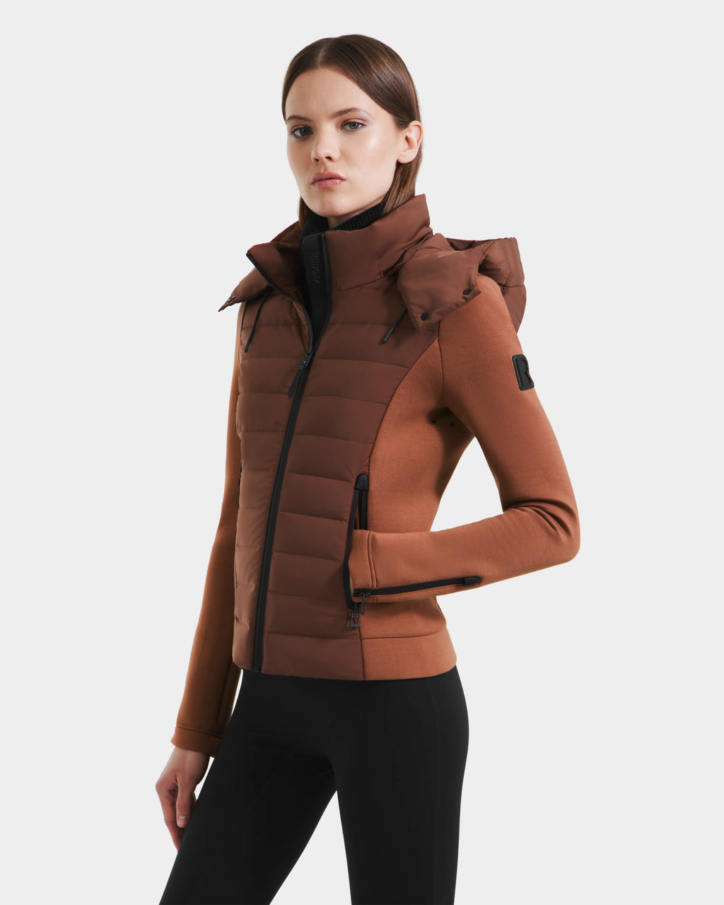 A woman wears the Rudsak MABS - 8125055 Rust, a brown fitted womens puffer jacket with a high collar and hood, paired with black leggings. This lightweight activewear uses premium RDS-certified down for warmth.