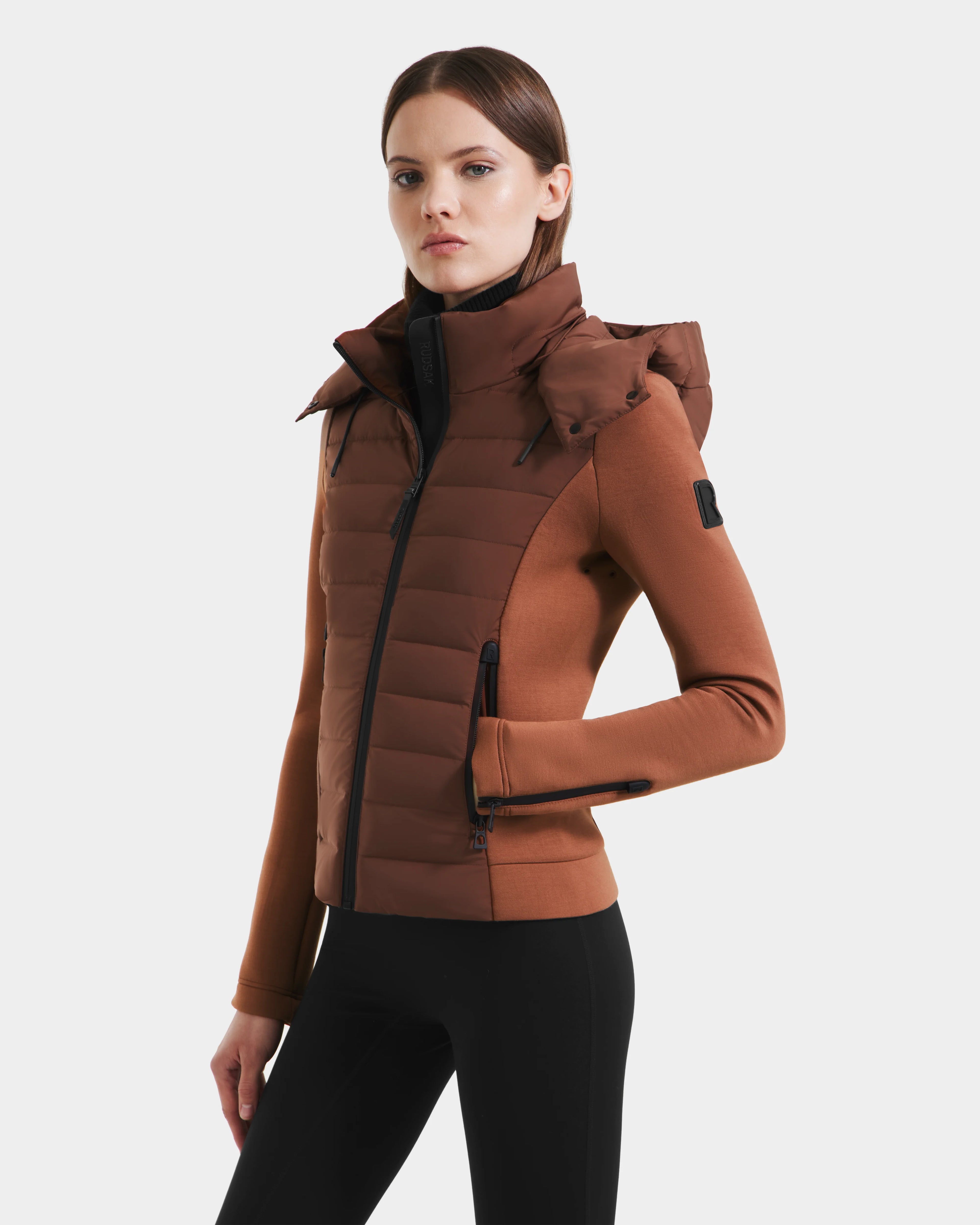 A woman wears the Rudsak MABS - 8125055 Rust, a brown fitted womens puffer jacket with a high collar and hood, paired with black leggings. This lightweight activewear uses premium RDS-certified down for warmth.