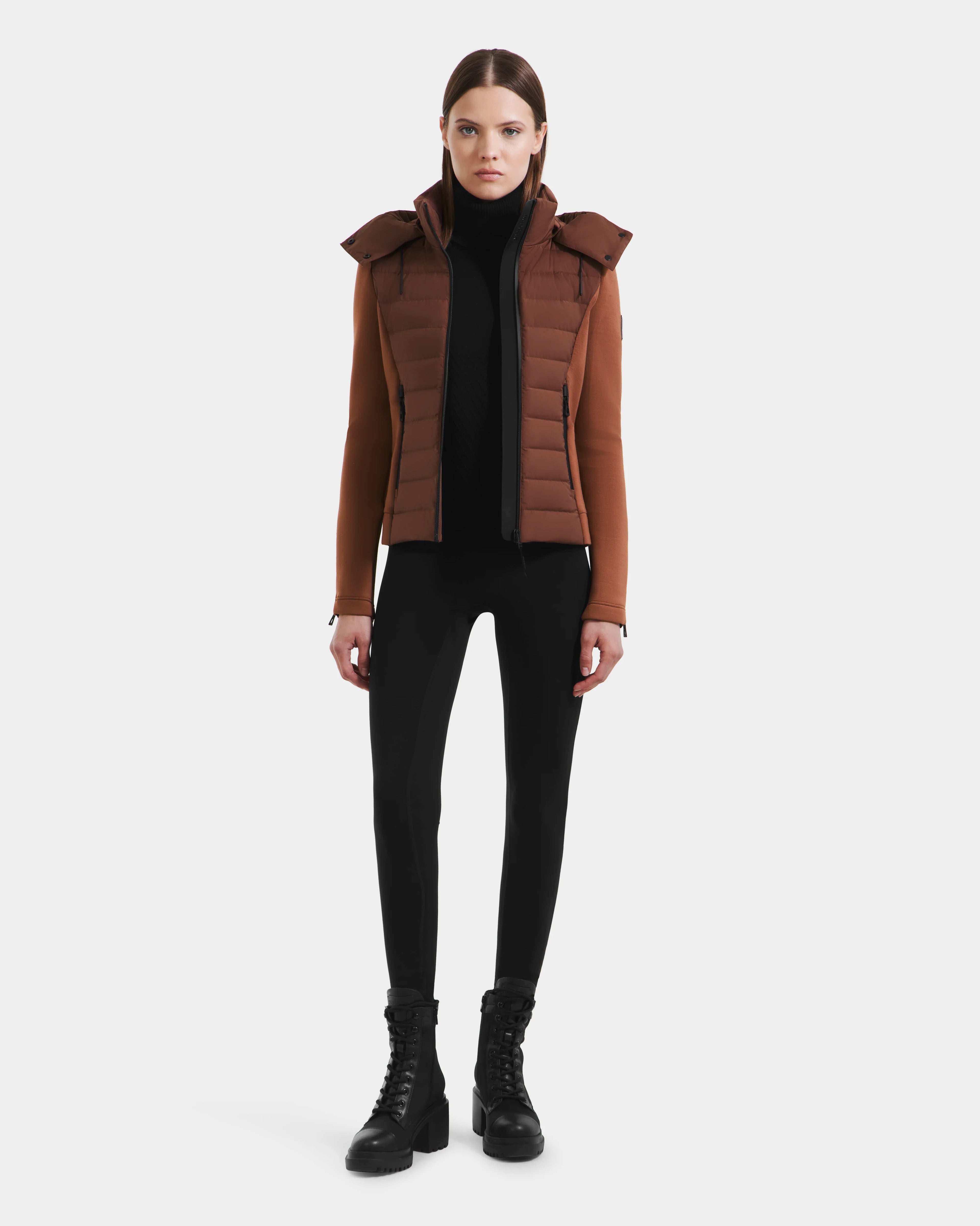 A woman faces forward against a white background, wearing the Rudsak MABS - 8125055 Rust women’s puffer jacket with a hood, paired with a black turtleneck, fitted pants, and lace-up boots.