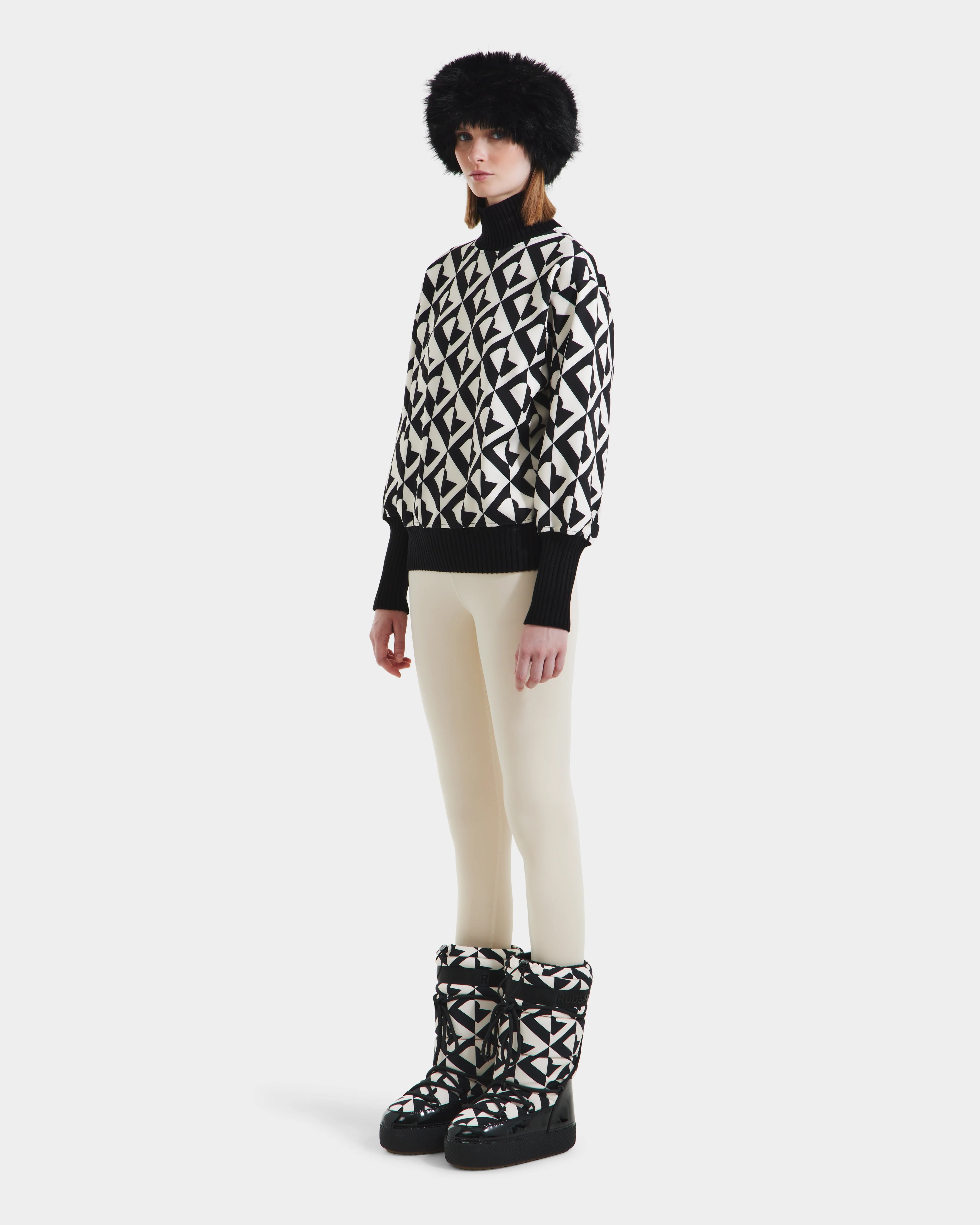 A person stands slightly to the side, wearing Rudsaks MAGGIE - 8525645 Vanilla Monogram winter boots, paired with a black fur hat, an alpine knit womens ski sweater with black-and-white patterns, and cream leggings against a white background.