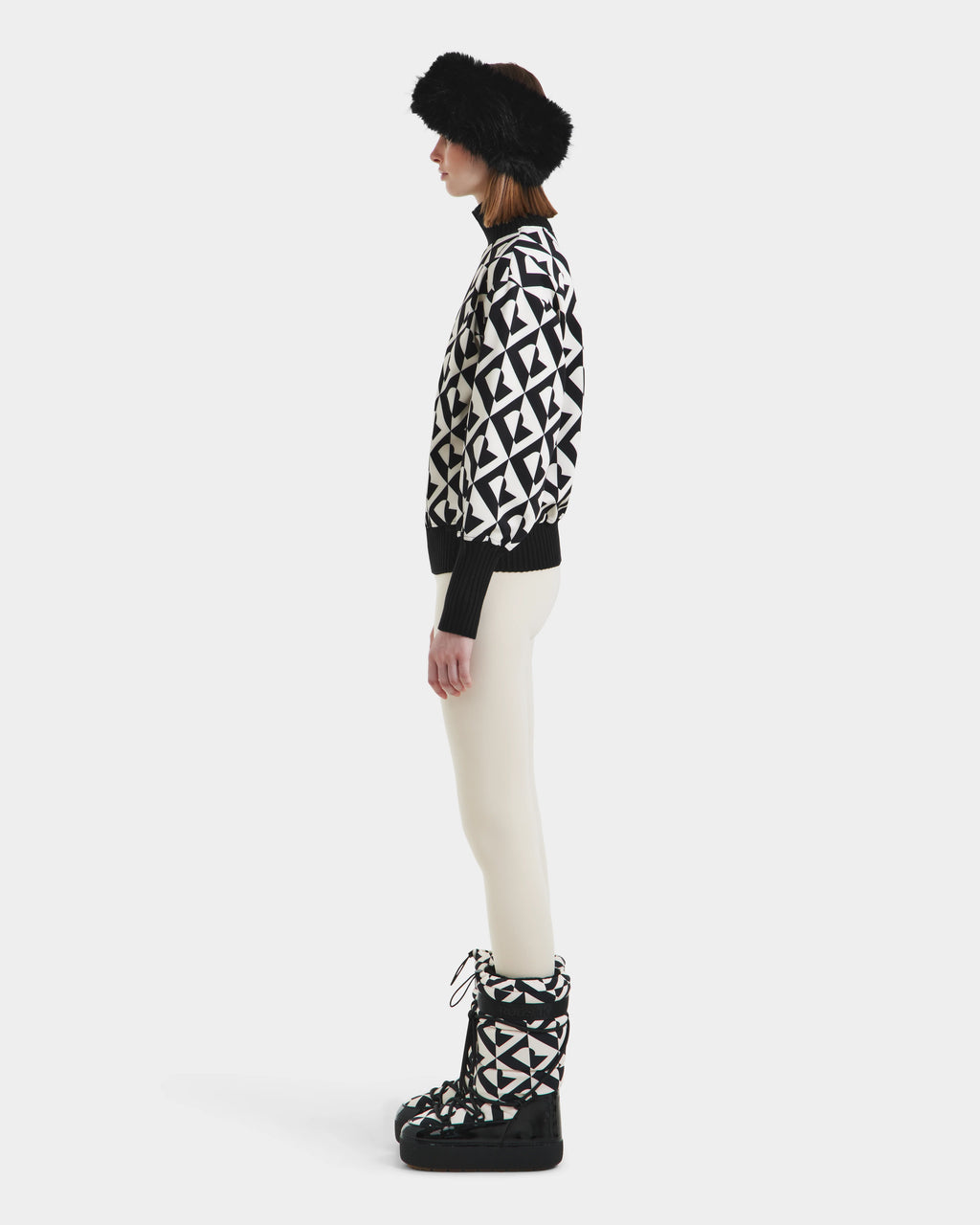 A person stands in profile against a plain white background, wearing Rudsaks MAGGIE - 8525645 Vanilla Monogram boots, paired with a black fur hat, a black and white geometric cotton ski sweater, and white pants.