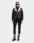 A woman in black sunglasses, the Rudsak MALA - 8125628 Black cropped shearling biker jacket, a black turtleneck, pants, and black lace-up boots with white fur lining stands against a plain white background.