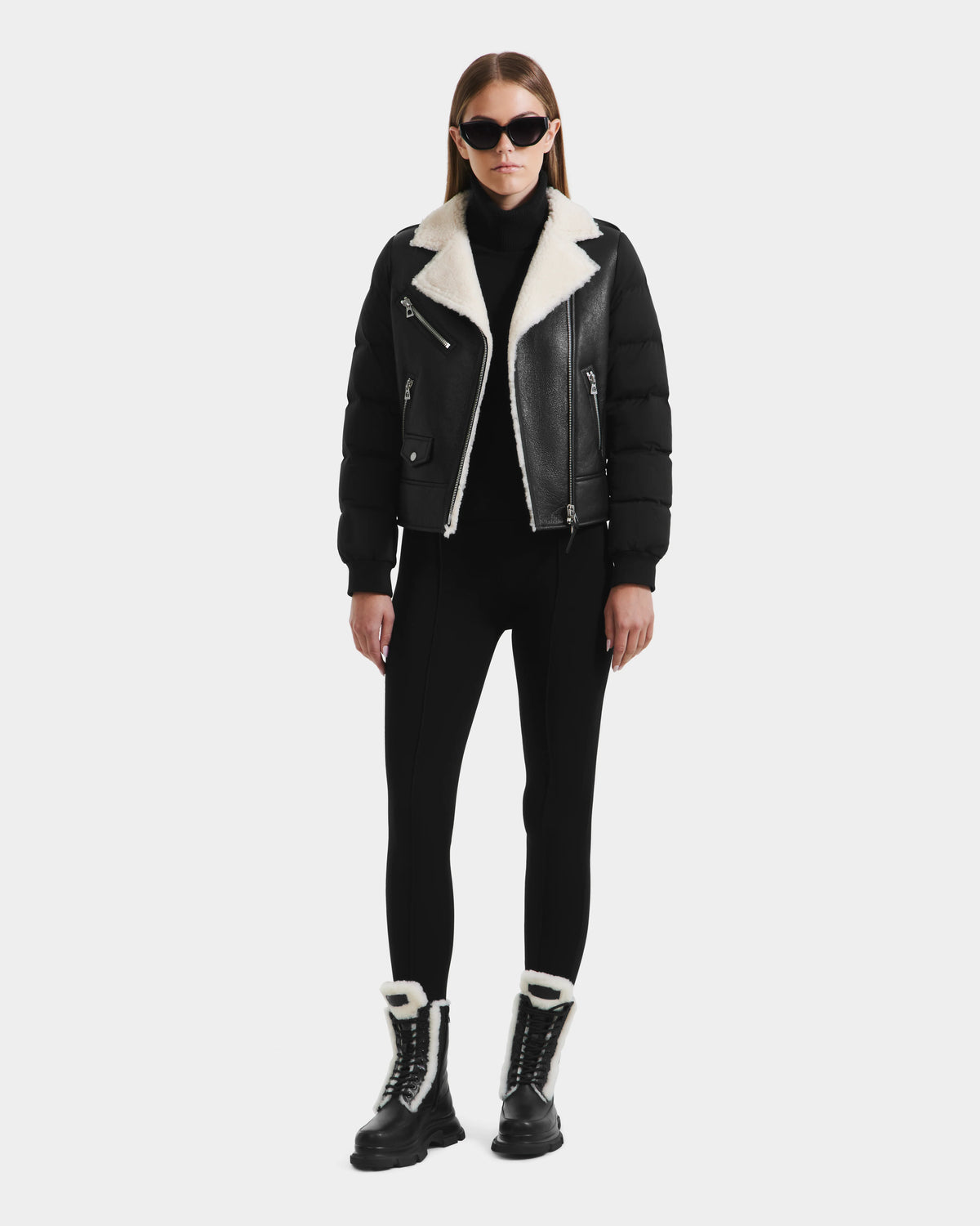 A woman in black sunglasses, the Rudsak MALA - 8125628 Black cropped shearling biker jacket, a black turtleneck, pants, and black lace-up boots with white fur lining stands against a plain white background.