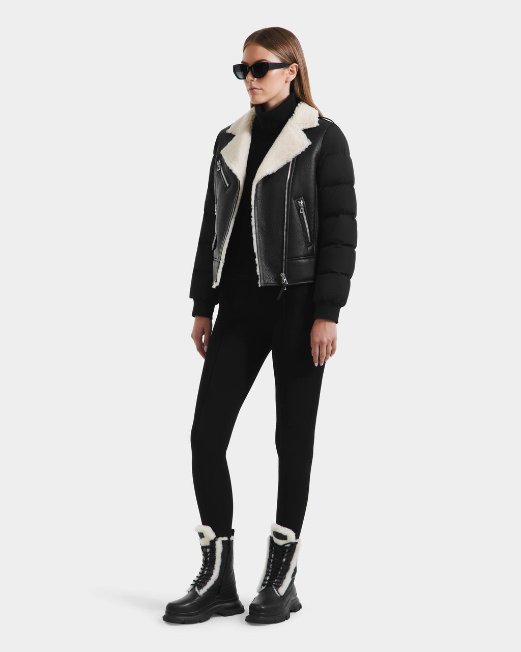 A woman in black sunglasses, the Rudsak MALA - 8125628 Black women’s shearling jacket with puffer sleeves, black leggings, and black lace-up boots with white shearling stands against a plain white background.