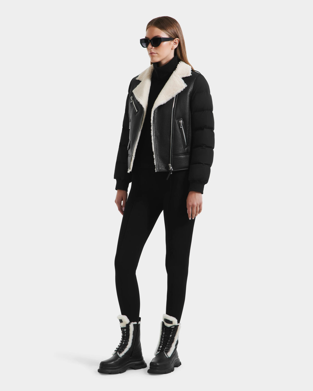 A woman in black sunglasses, the Rudsak MALA - 8125628 Black women’s shearling jacket with puffer sleeves, black leggings, and black lace-up boots with white shearling stands against a plain white background.