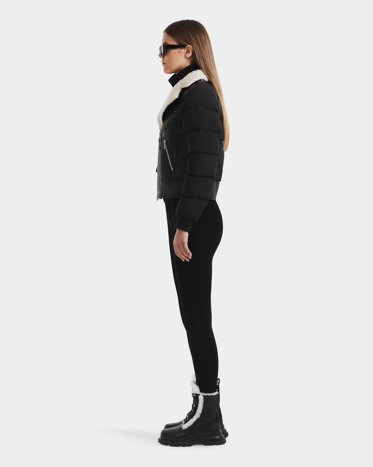 A woman in profile wears black sunglasses, the Rudsak MALA - 8125628 Black puffer jacket with a white shearling collar, black leggings, and black boots with white trim against a plain white background.
