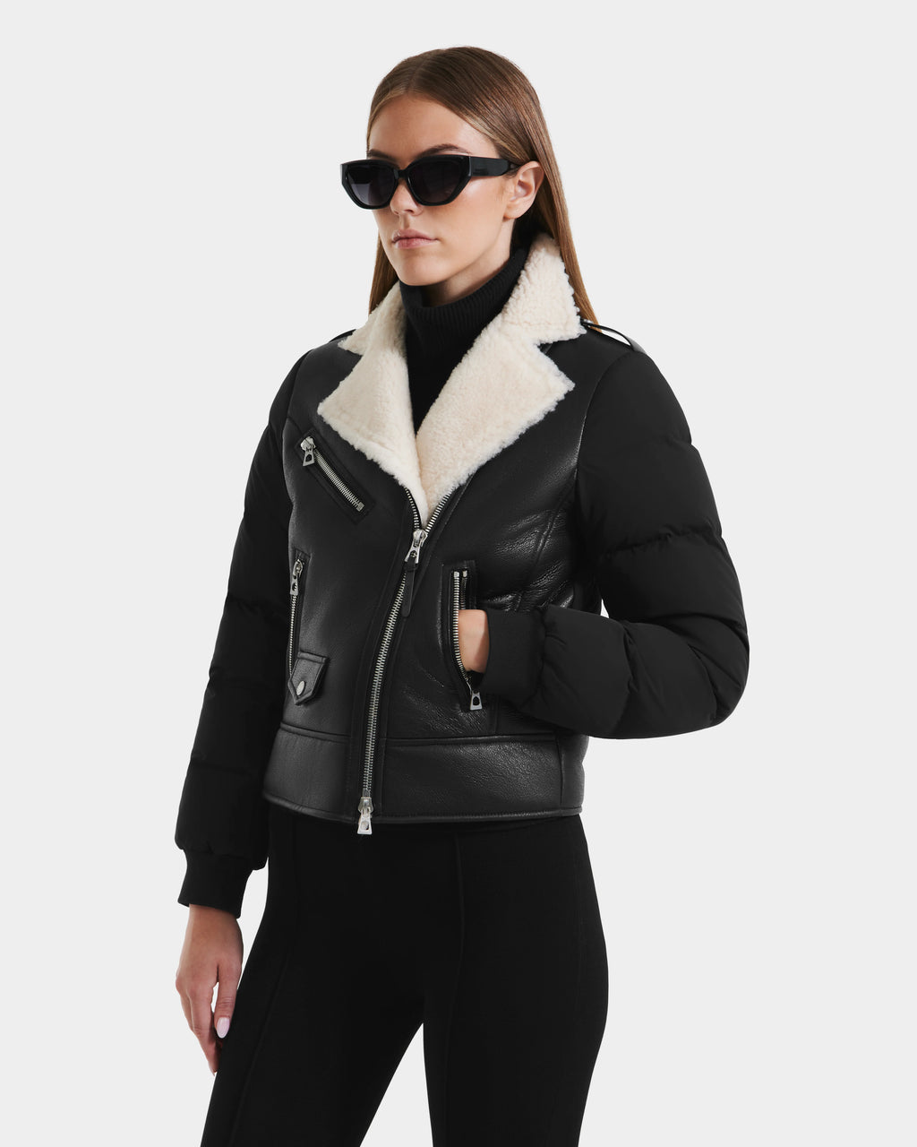 A woman wearing black sunglasses and black pants models the Rudsak MALA - 8125628 Black women’s shearling jacket, featuring a white collar and cropped silhouette, standing against a plain white background.