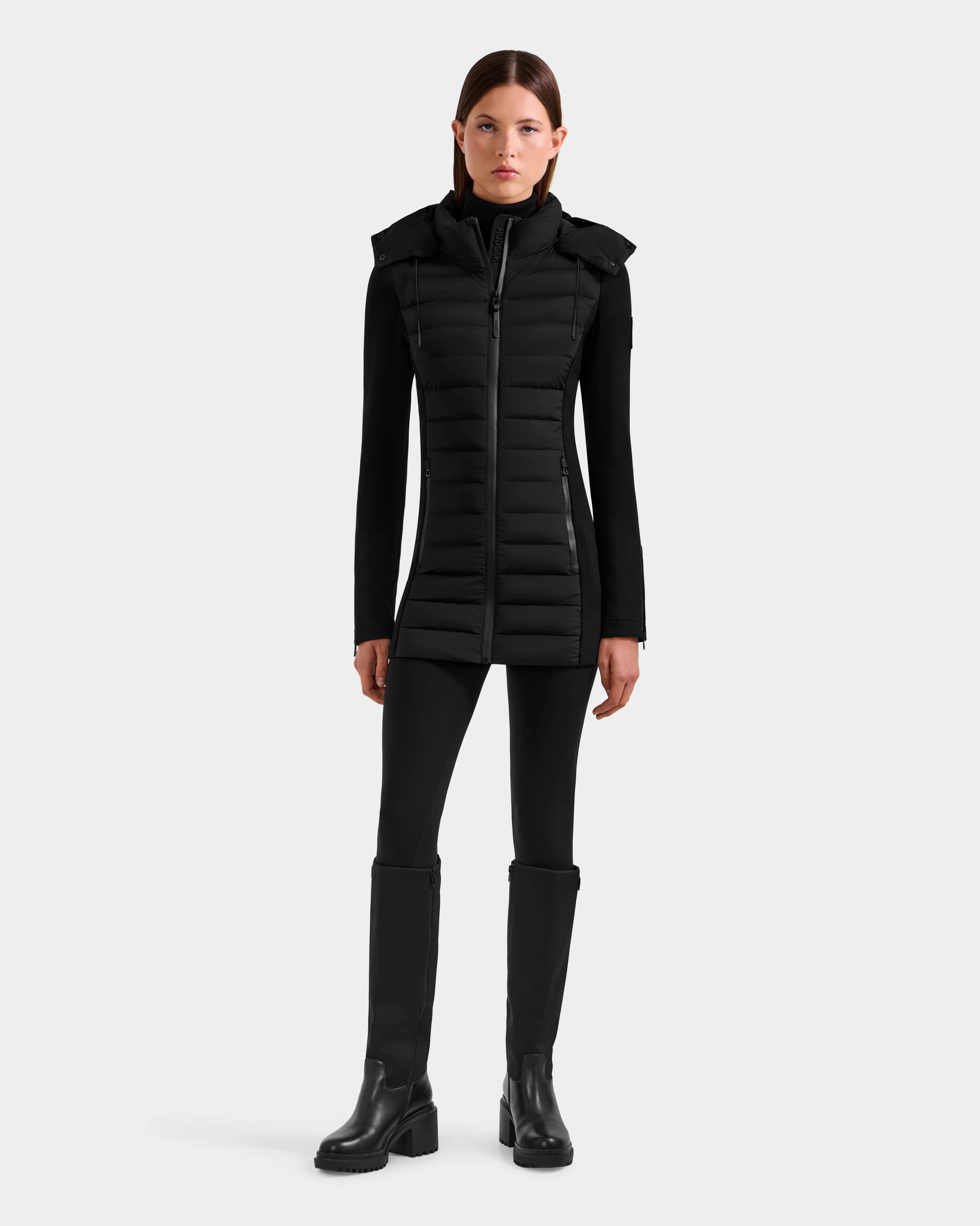 A woman faces forward in the Rudsak MARRY M - L125519 Black women's mid-length hybrid coat, layered over a black top, pants, and knee-high boots against a plain white background.