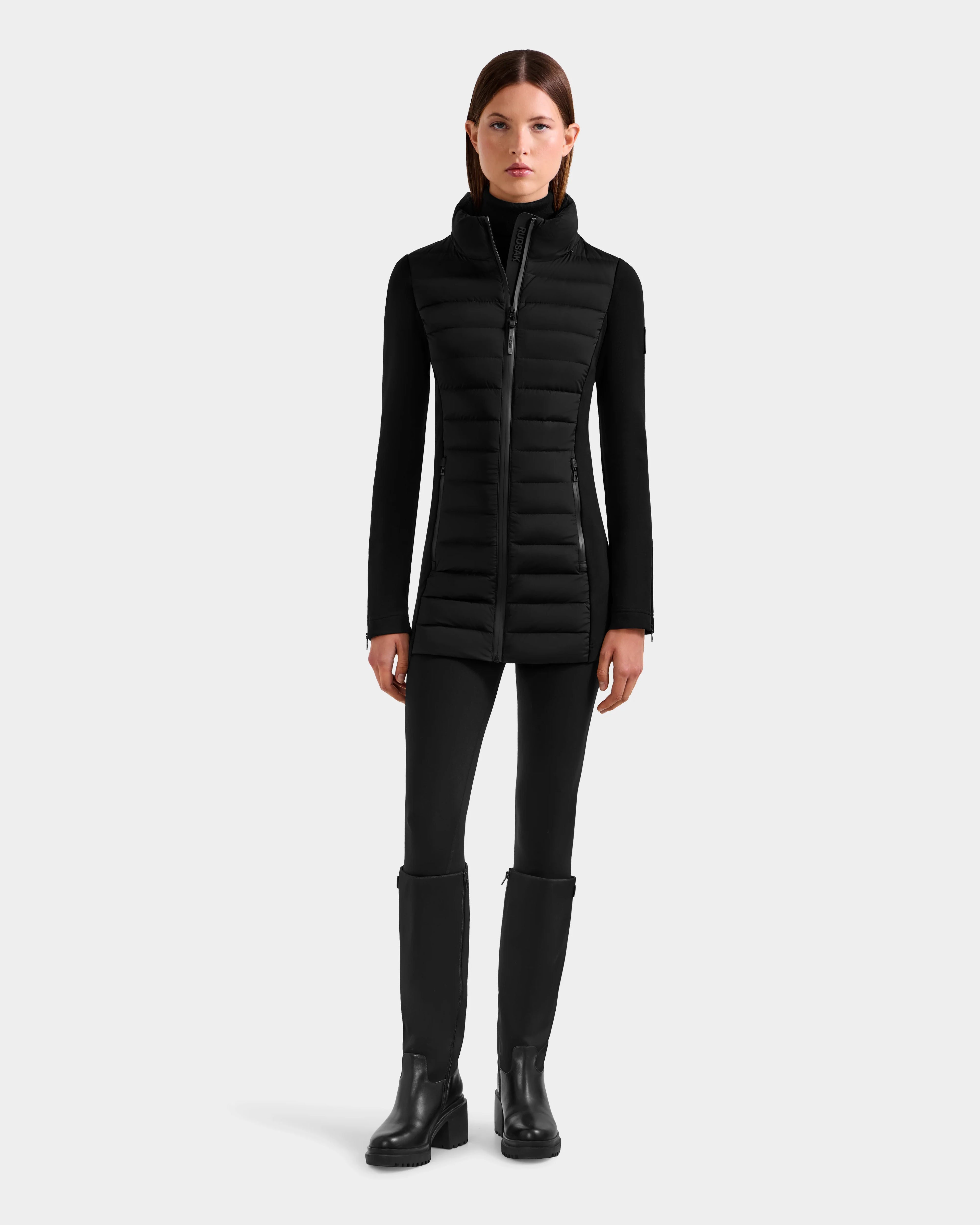 A woman stands facing forward in a Rudsak MARRY M - L125519 black mid-length hybrid coat, paired with black pants and knee-high boots, against a plain light gray background.