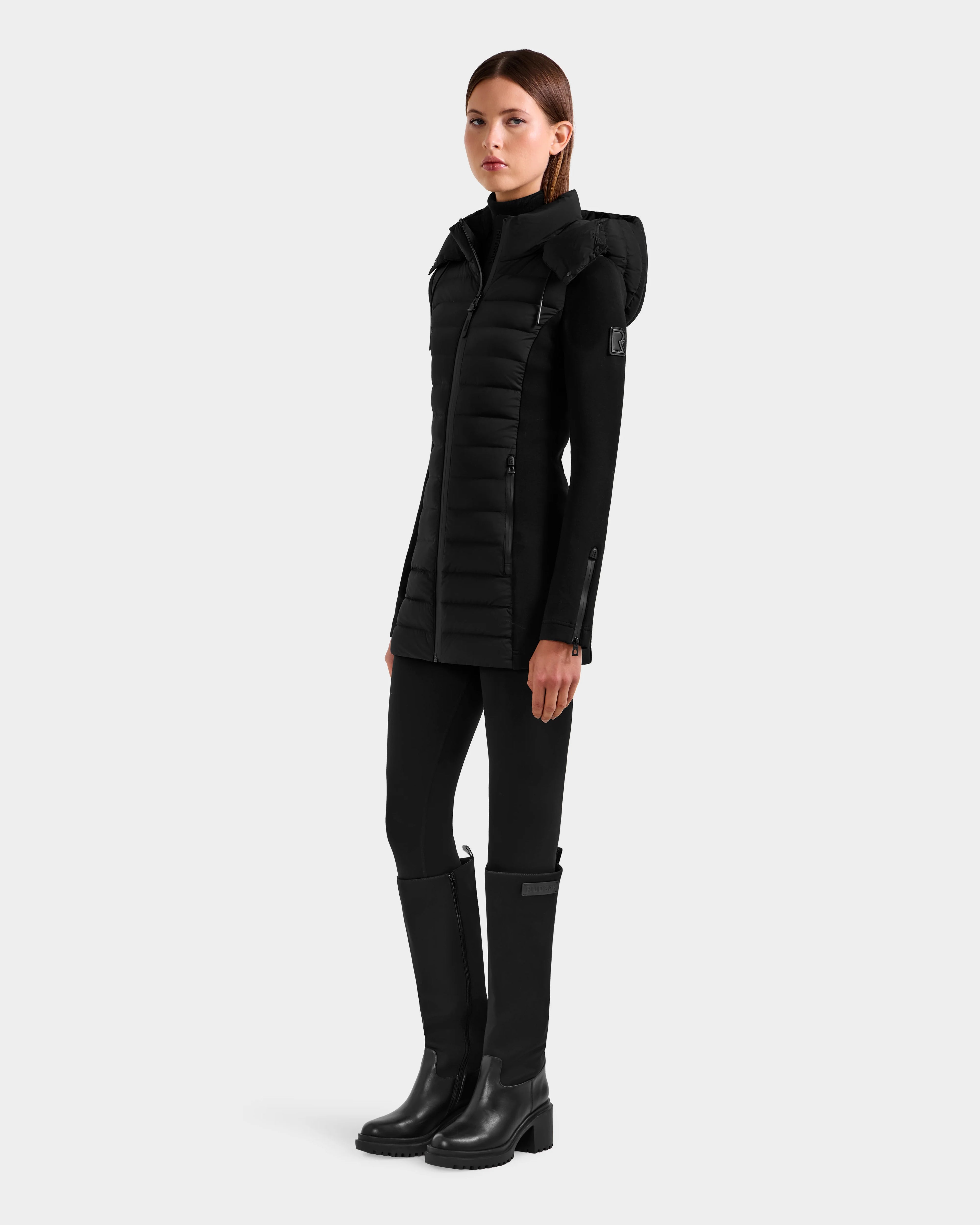 A woman faces slightly left, wearing the Rudsak MARRY M - L125519 Black hooded puffer vest over a black turtleneck, slim black pants, and knee-high boots against a light gray background.