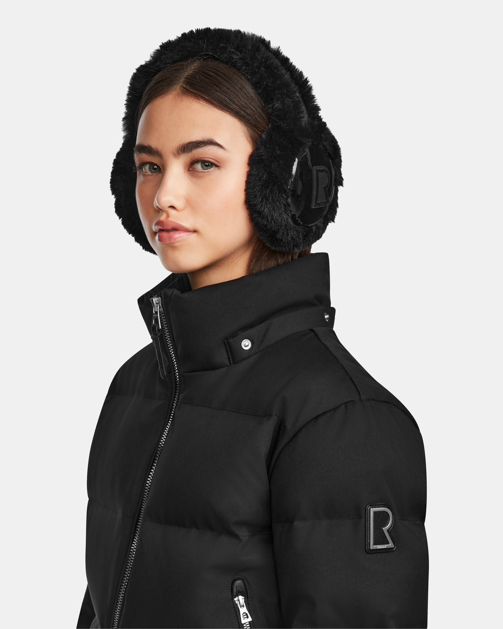 A young woman wears Rudsak's MAUDE Q-M - L124516 Black puffer jacket with a high collar and black faux fur earmuffs, looking confidently at the camera against a plain white background.