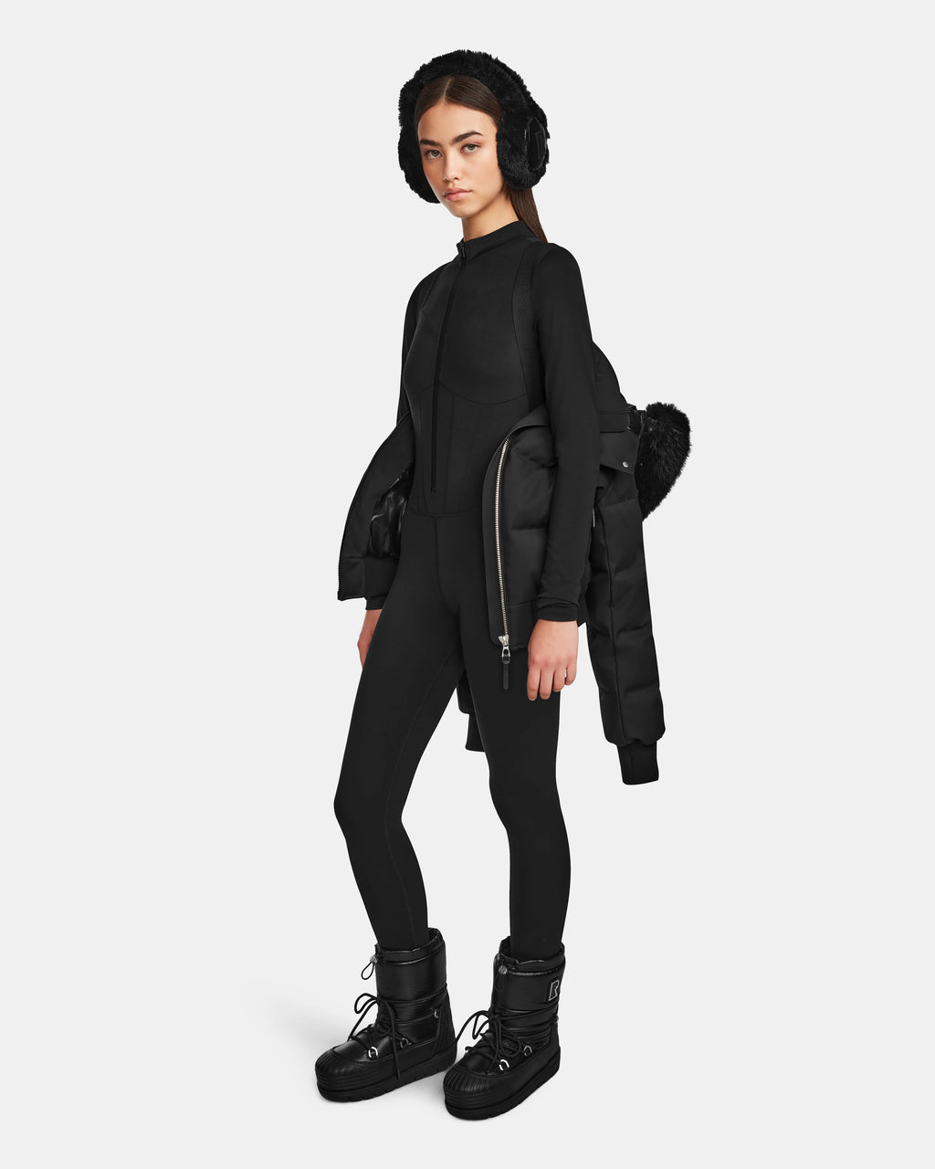A woman wears the Rudsak MAUDE Q-M - L124516 Black quilted down bomber over one shoulder, styled with black earmuffs, a fitted long-sleeve top, leggings, and chunky boots against a plain light background.