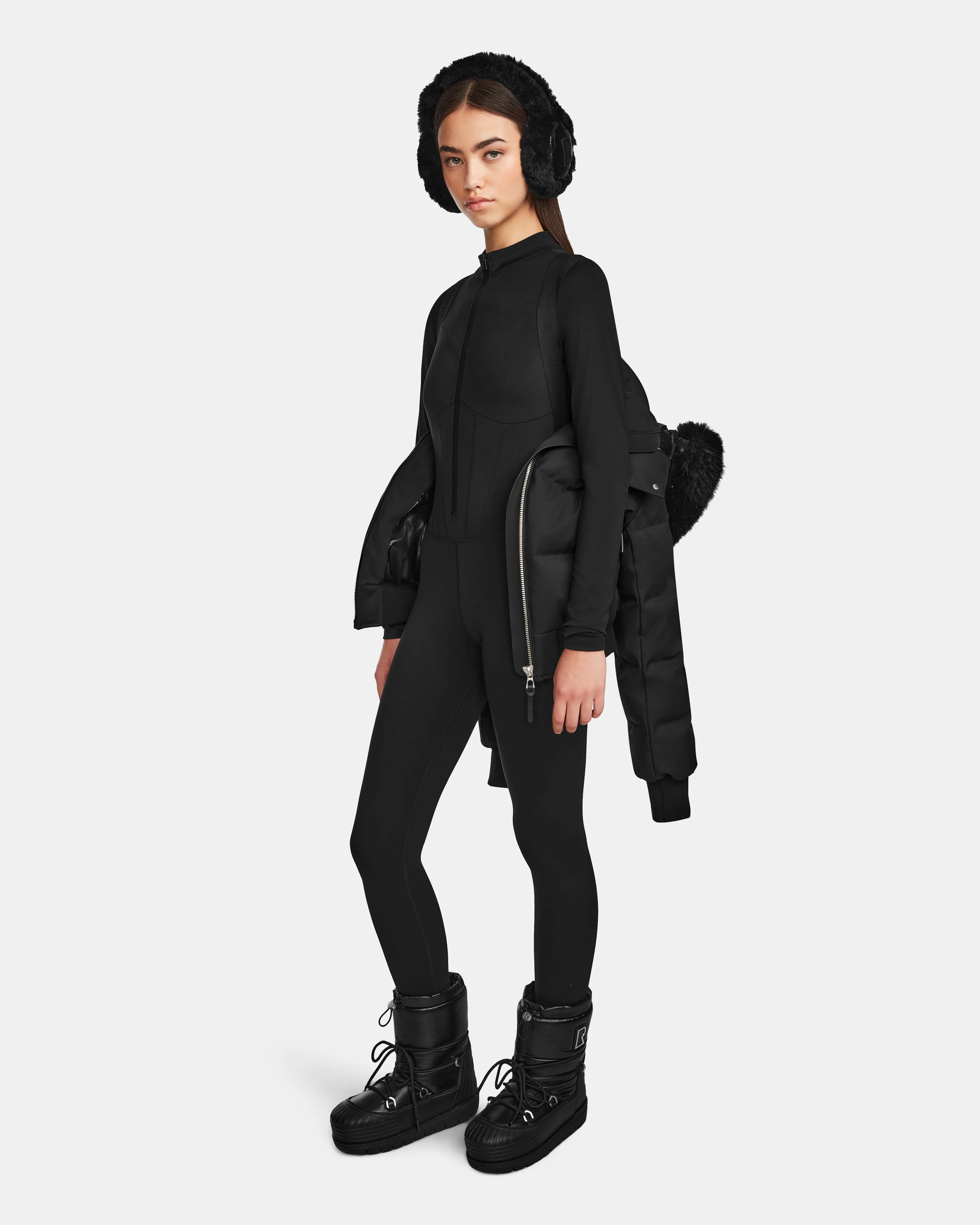 A woman wears the Rudsak MAUDE Q-M - L124516 Black quilted down bomber over one shoulder, styled with black earmuffs, a fitted long-sleeve top, leggings, and chunky boots against a plain light background.