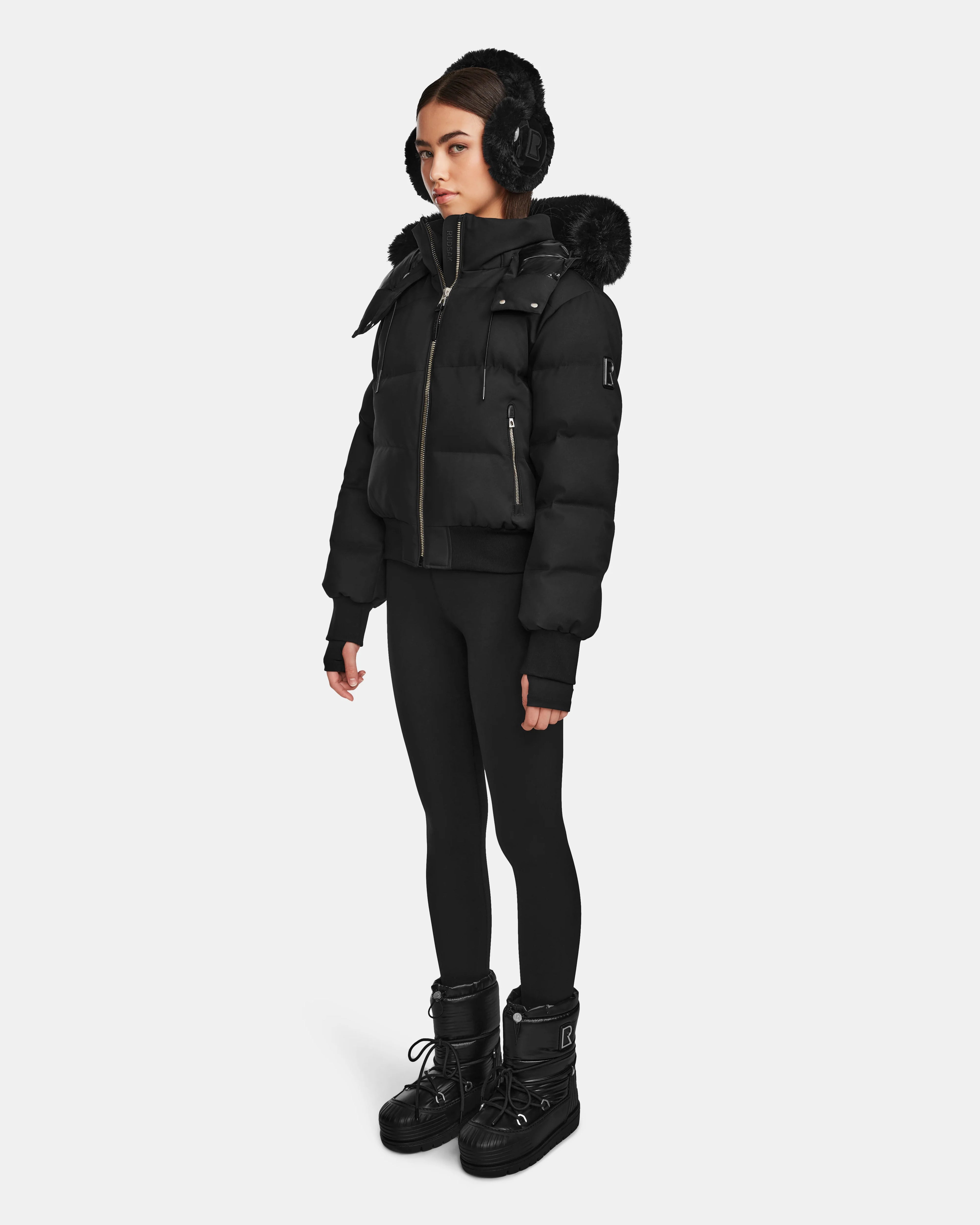 A woman stands against a white background wearing the Rudsak MAUDE Q-M - L124516 Black quilted down bomber jacket with a fur-trimmed hood, black earmuffs, black leggings, and black winter boots.
