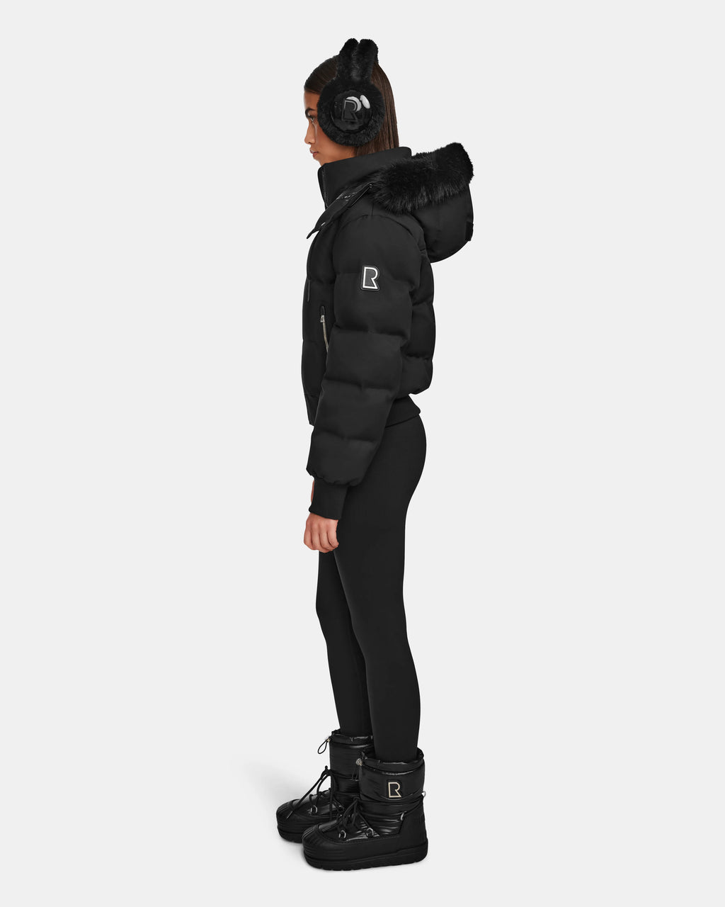 A woman stands in profile wearing Rudsak’s MAUDE Q-M - L124516 Black quilted down bomber with fur trim, black earmuffs, leggings, and winter boots. The warm, stylish outfit is perfect for cold weather against a plain white background.