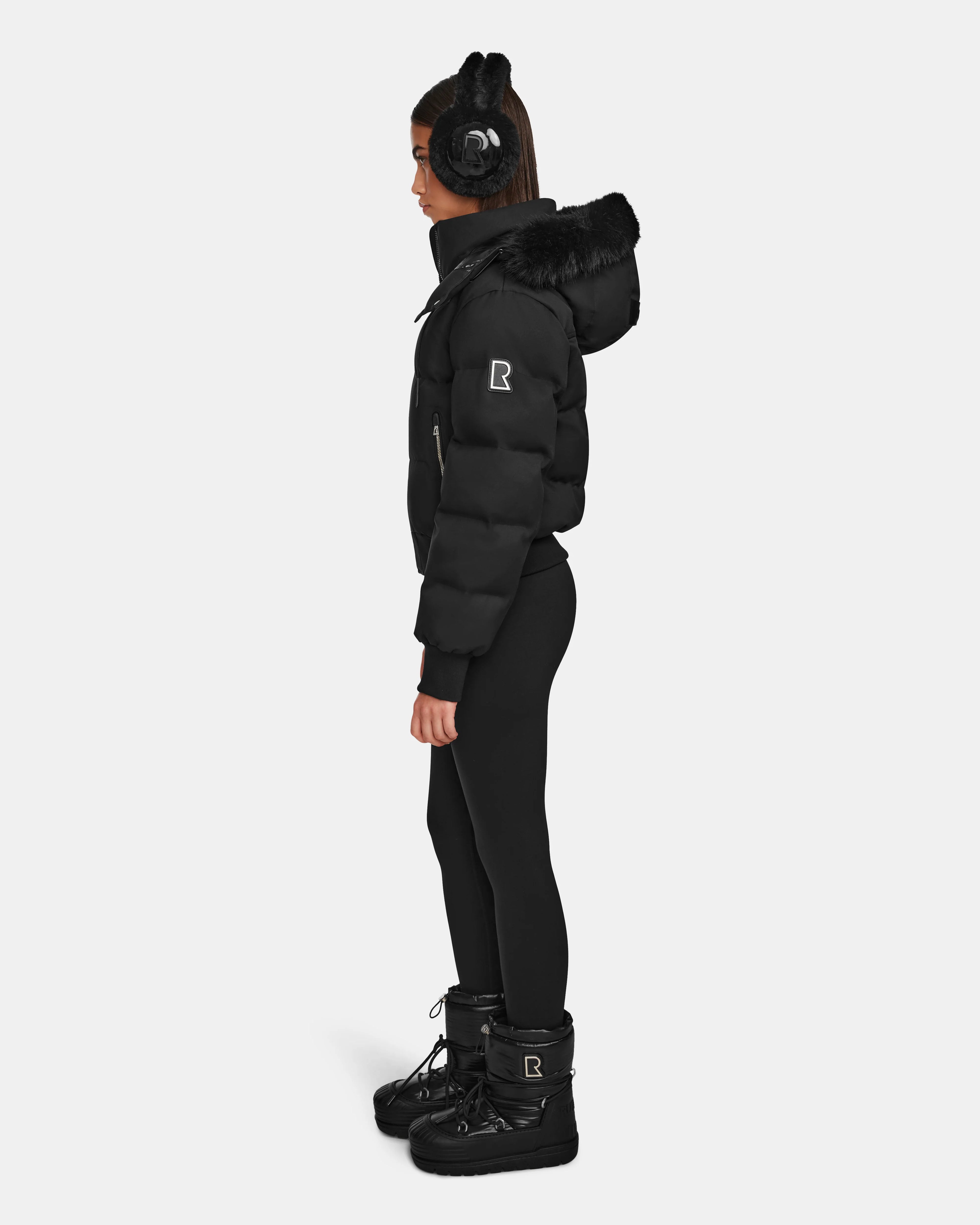 A woman stands in profile wearing Rudsak’s MAUDE Q-M - L124516 Black quilted down bomber with fur trim, black earmuffs, leggings, and winter boots. The warm, stylish outfit is perfect for cold weather against a plain white background.