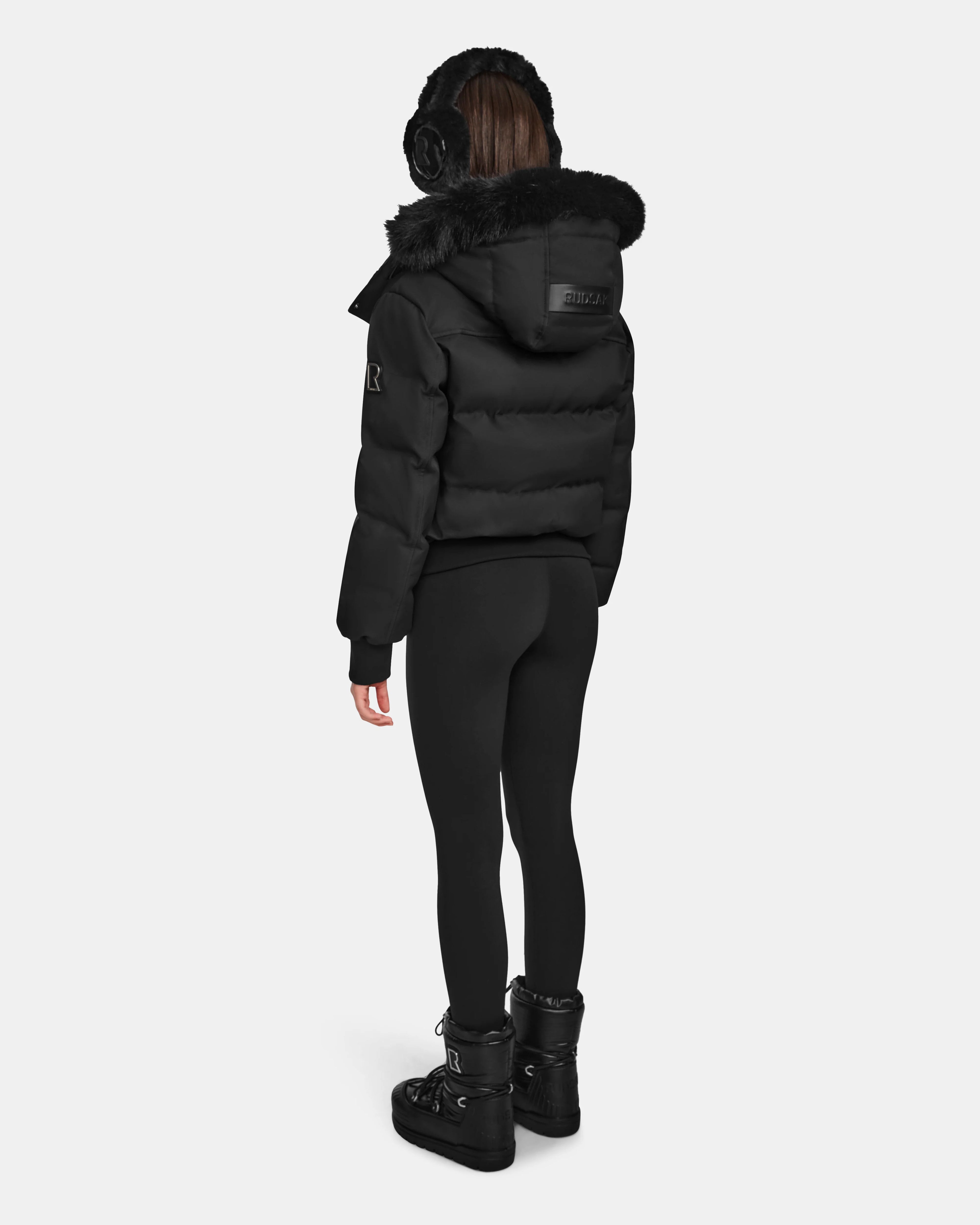 A person stands facing away, wearing Rudsak's MAUDE Q-M - L124516 Black women’s quilted down bomber puffer jacket with fur trim, black earmuffs, leggings, and winter boots against a plain white background.
