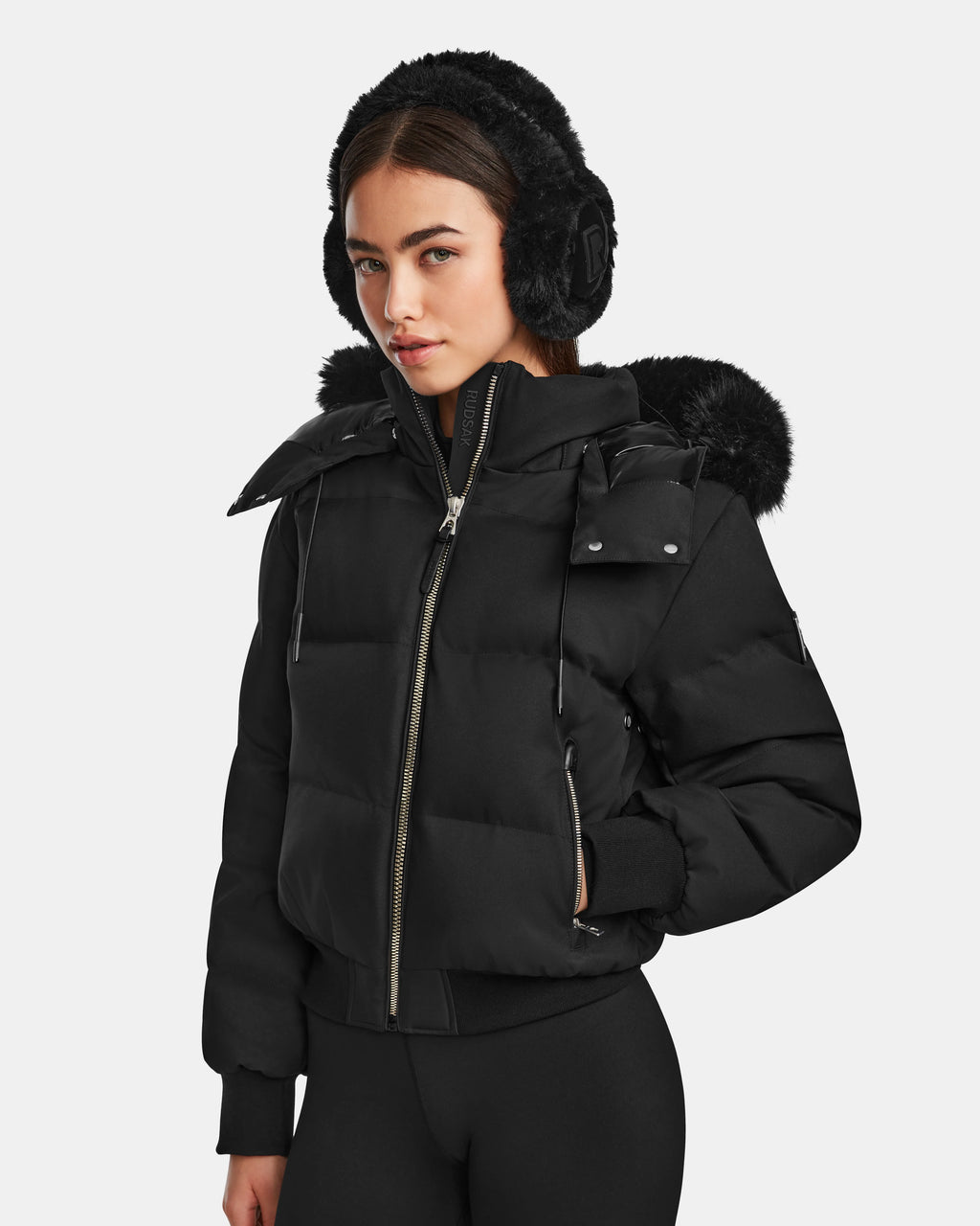 A woman with long dark hair wears the Rudsak MAUDE Q-M - L124516 Black down jacket featuring a fur-trimmed hood and black earmuffs, standing against a plain light background and looking slightly to the side.