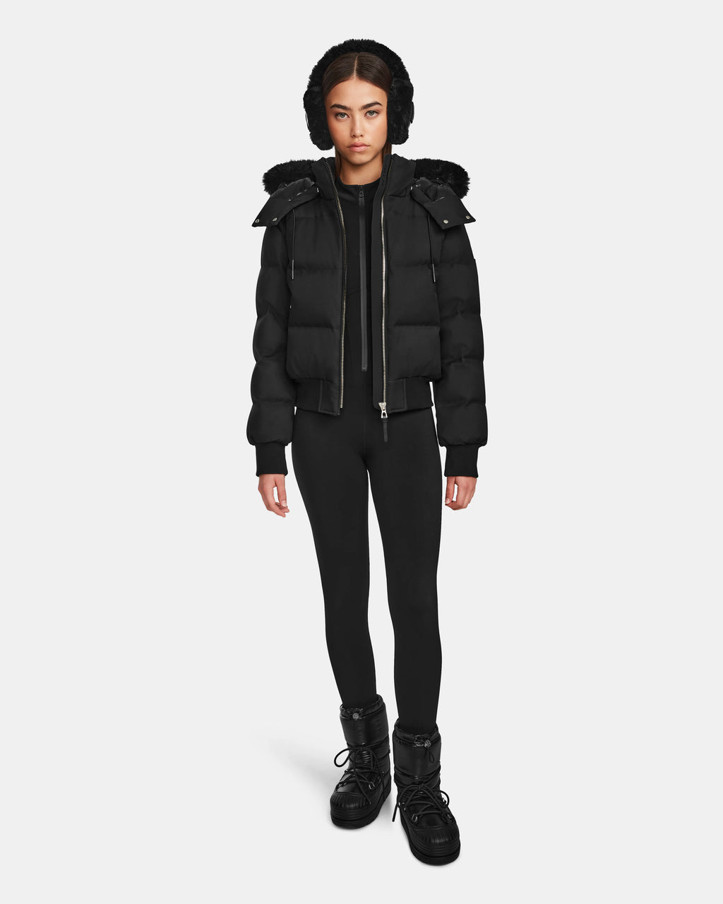 A woman faces forward, wearing the Rudsak MAUDE Q-M - L124516 Black quilted down bomber with a hood, paired with black leggings and winter boots against a plain white background.