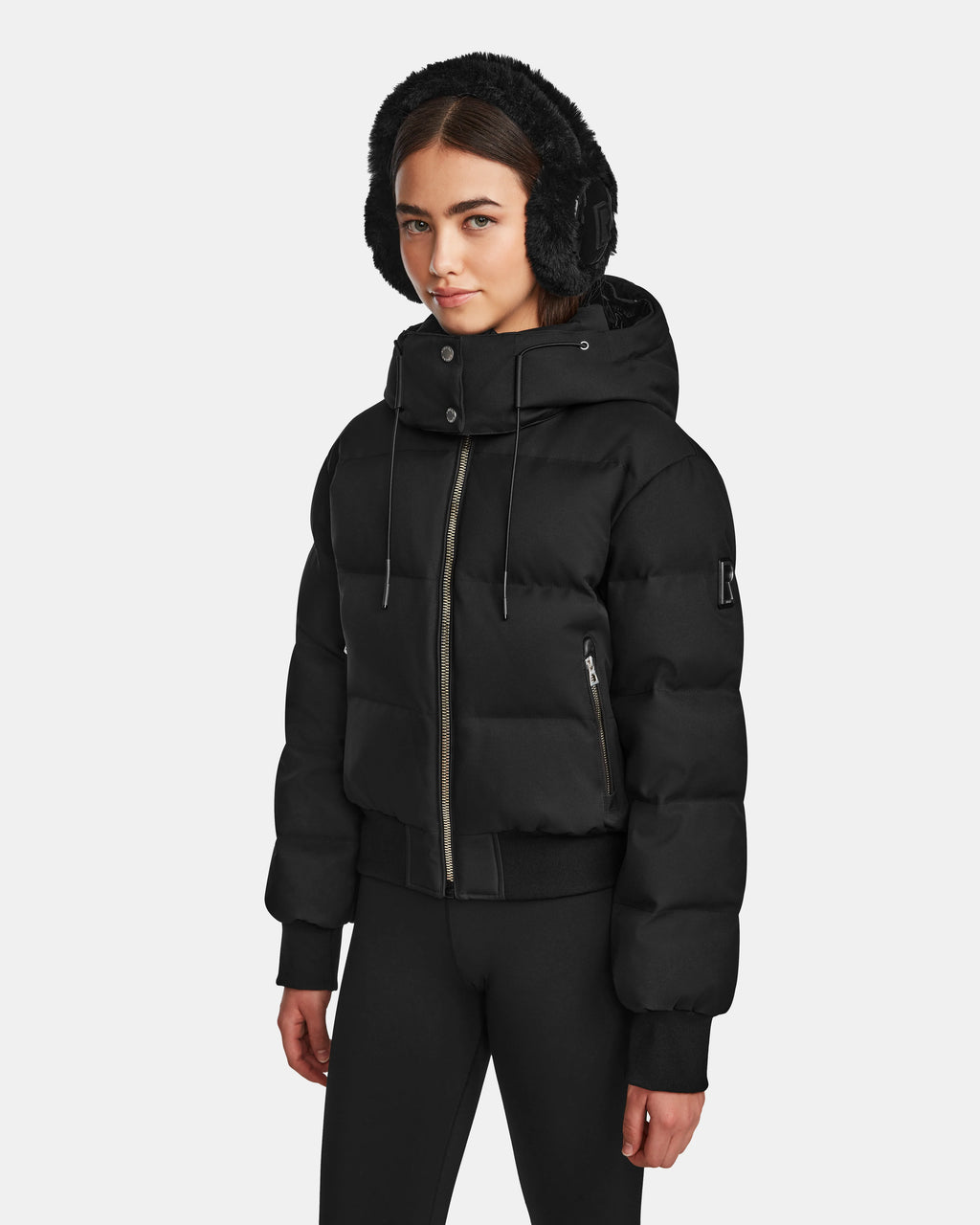A young woman with long dark hair wears the Rudsak MAUDE Q-M - L124516 Black quilted down bomber with a hood and black earmuffs. Dressed in black leggings, she stands against a plain white background, ready for winter.
