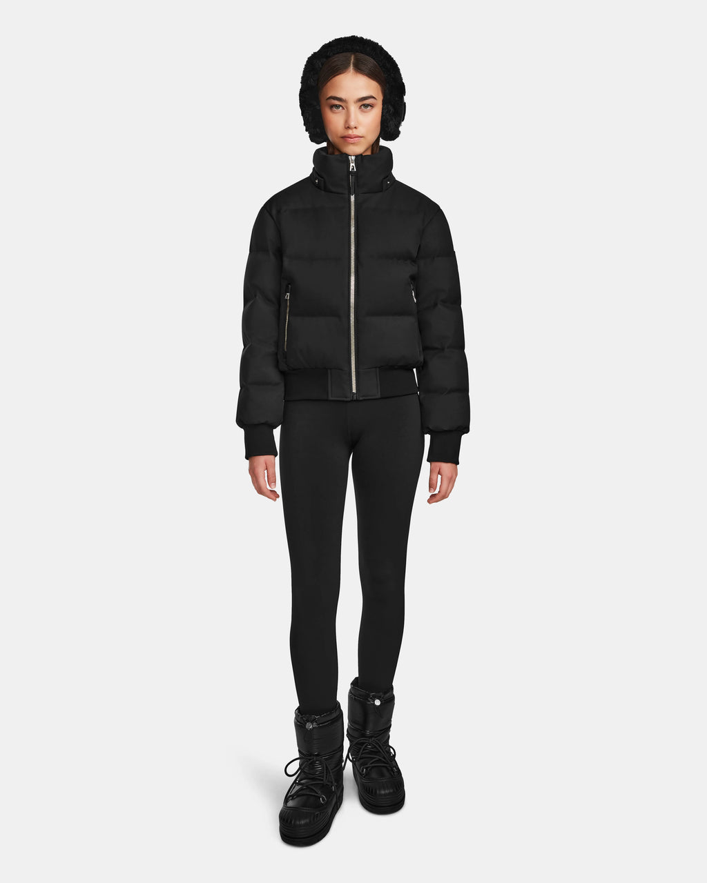 A person stands facing forward, wearing the Rudsak MAUDE Q-M - L124516 Black women's quilted down bomber with a hood, black leggings, and black winter boots, against a plain white background.