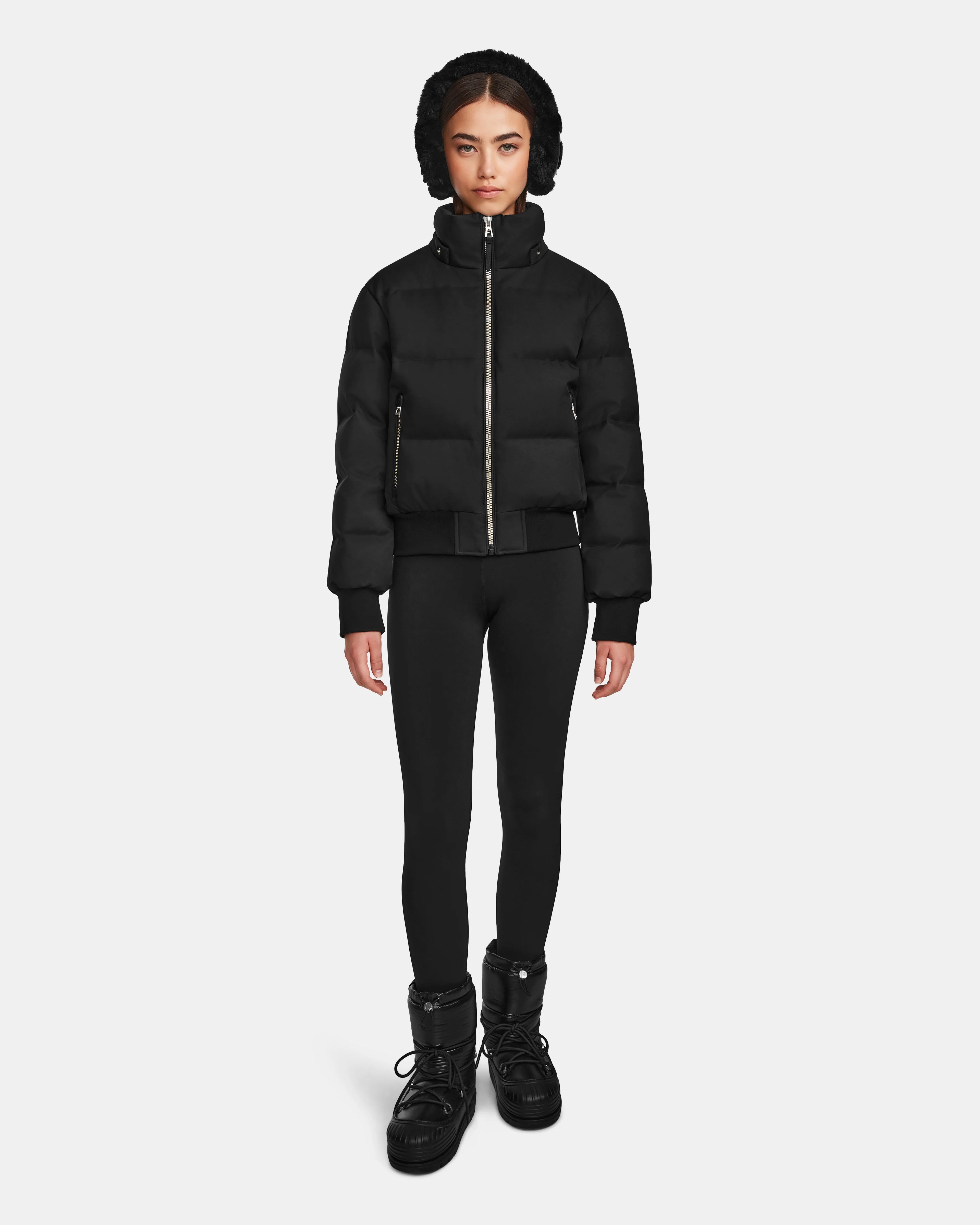 A person stands facing forward, wearing the Rudsak MAUDE Q-M - L124516 Black women's quilted down bomber with a hood, black leggings, and black winter boots, against a plain white background.