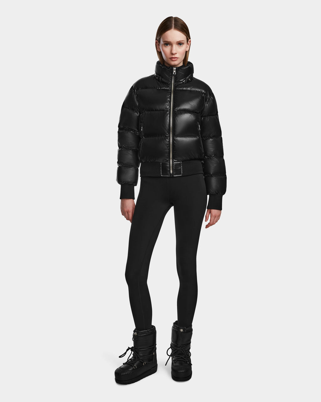 A woman stands against a plain background wearing the Rudsak MAUDE Q-S - L124553 Black down jacket, black leggings, and winter boots. She has straight hair, a neutral expression, and her hands are relaxed by her sides.