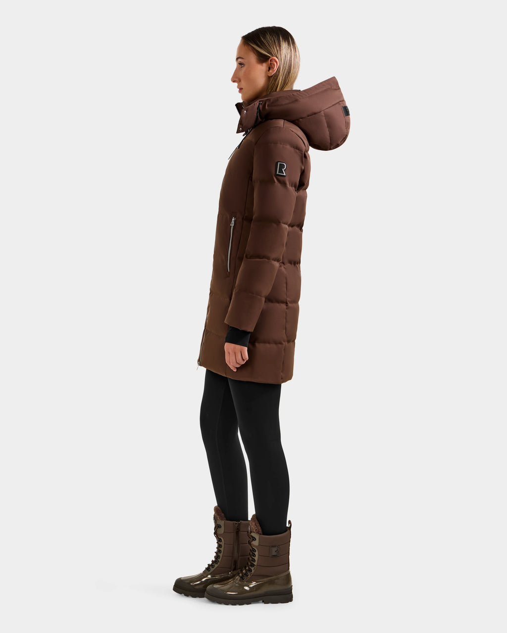 A woman in profile wears the Rudsak MIRIAM - L123544 Mocha weather-resistant hooded puffer coat, paired with black leggings and brown winter boots, standing against a plain white background.
