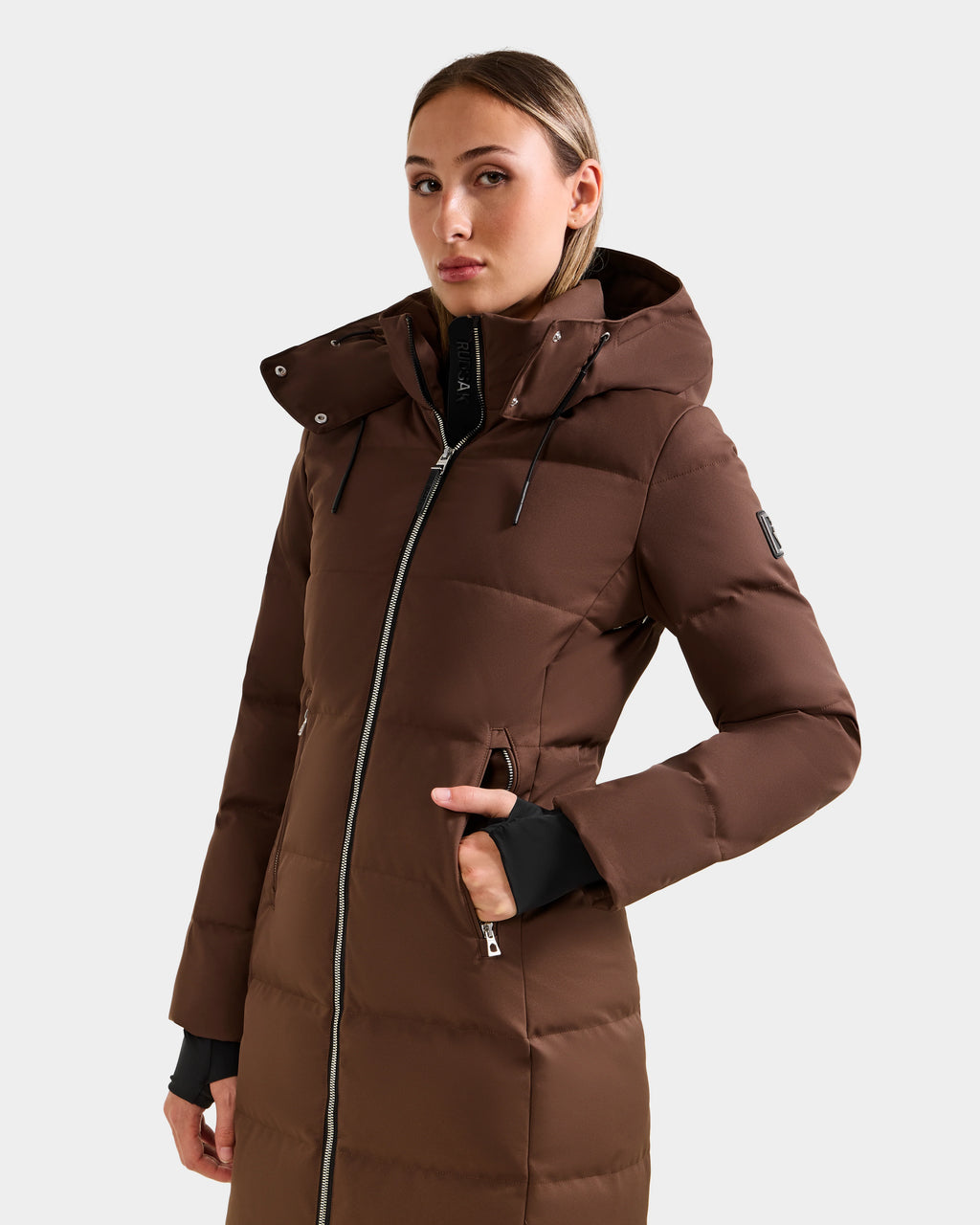 Wearing the Rudsak MIRIAM - L123544 Mocha women’s down puffer with hood, front zipper, and zippered pockets, a person stands against a light background—a stylish and cozy winter essential.