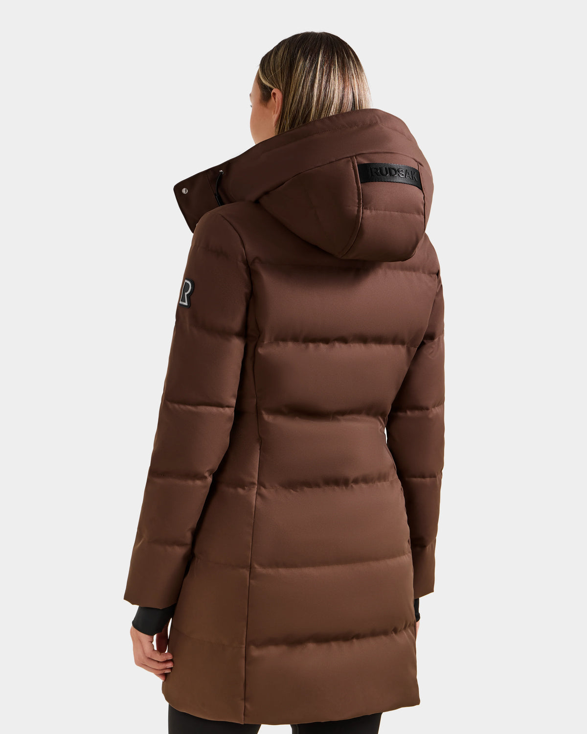 A person is seen from the back wearing the Rudsak MIRIAM - L123544 Mocha women's down puffer coat, featuring a hood, horizontal quilting, an upper-arm patch, and a weather-resistant shell against a plain light background.
